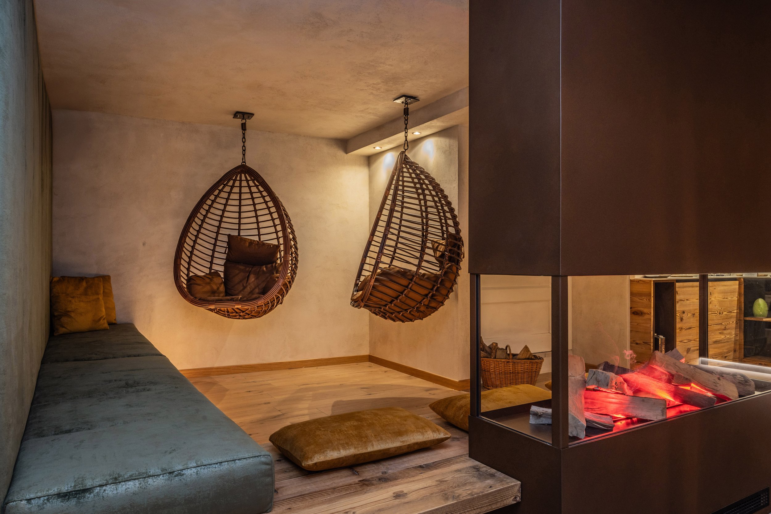 Interno di una stanza con sedute di legno e cuscini, due poltrone di vimini sospese dal soffitto e un camino con braci e fiamma.
