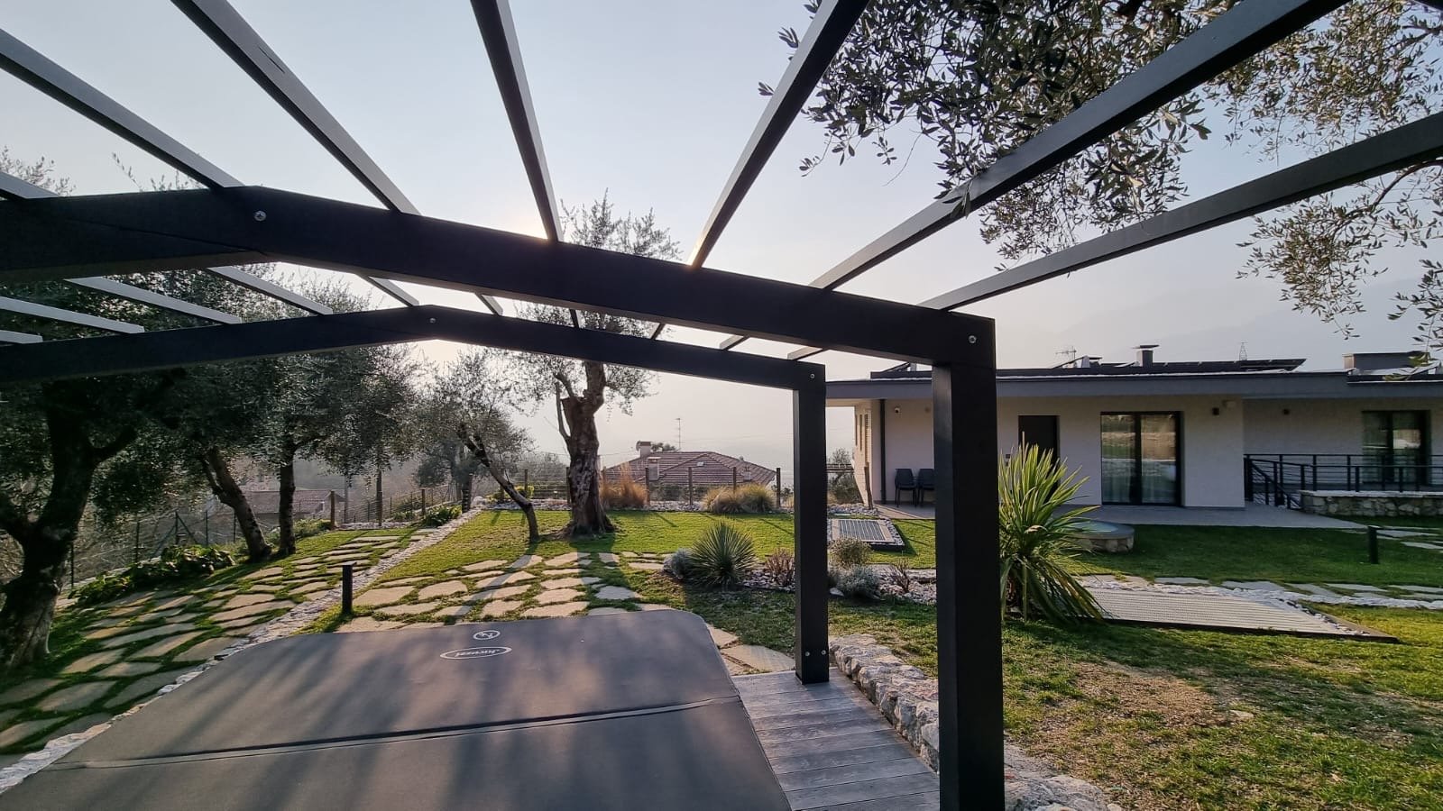 Giardino con panchina sotto una struttura in metallo, alberi, verifica, casa moderna con balcone e aree verdi.