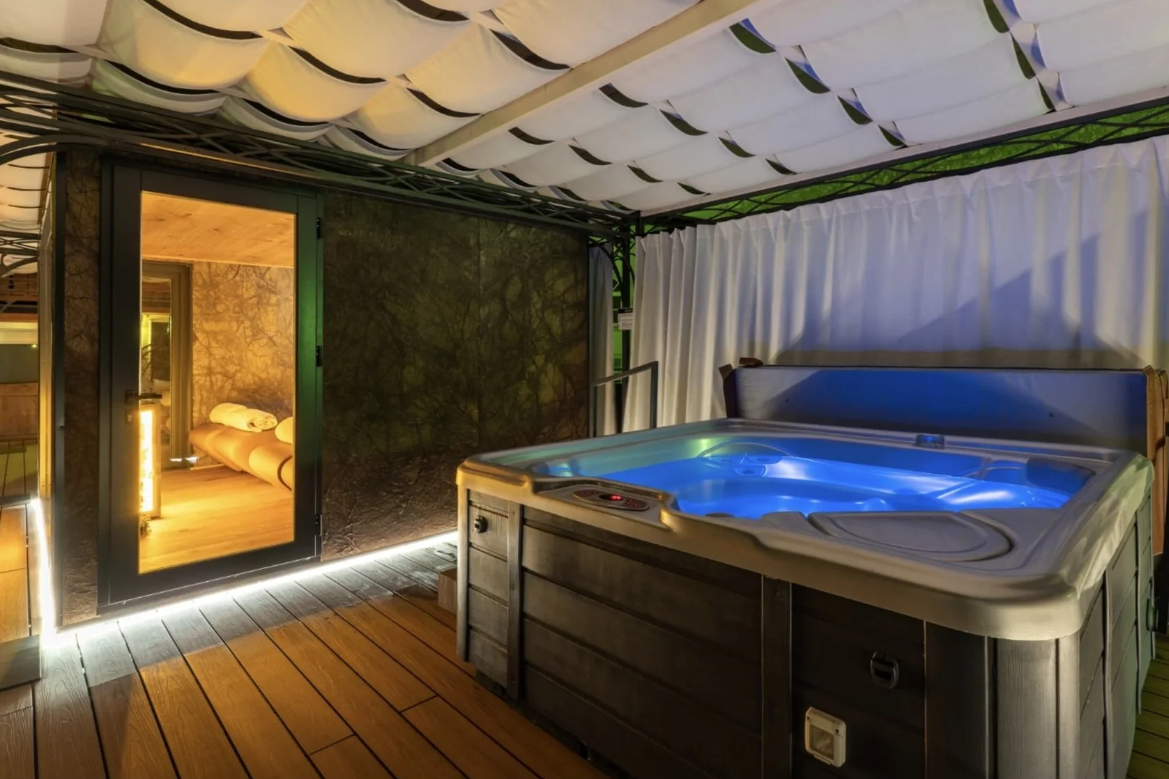 Spa con jacuzzi con illuminazione blu, stanza riscaldata visibile attraverso una porta con vetro, pavimento in legno, tende bianche e tetto rivestito con assi di legno.