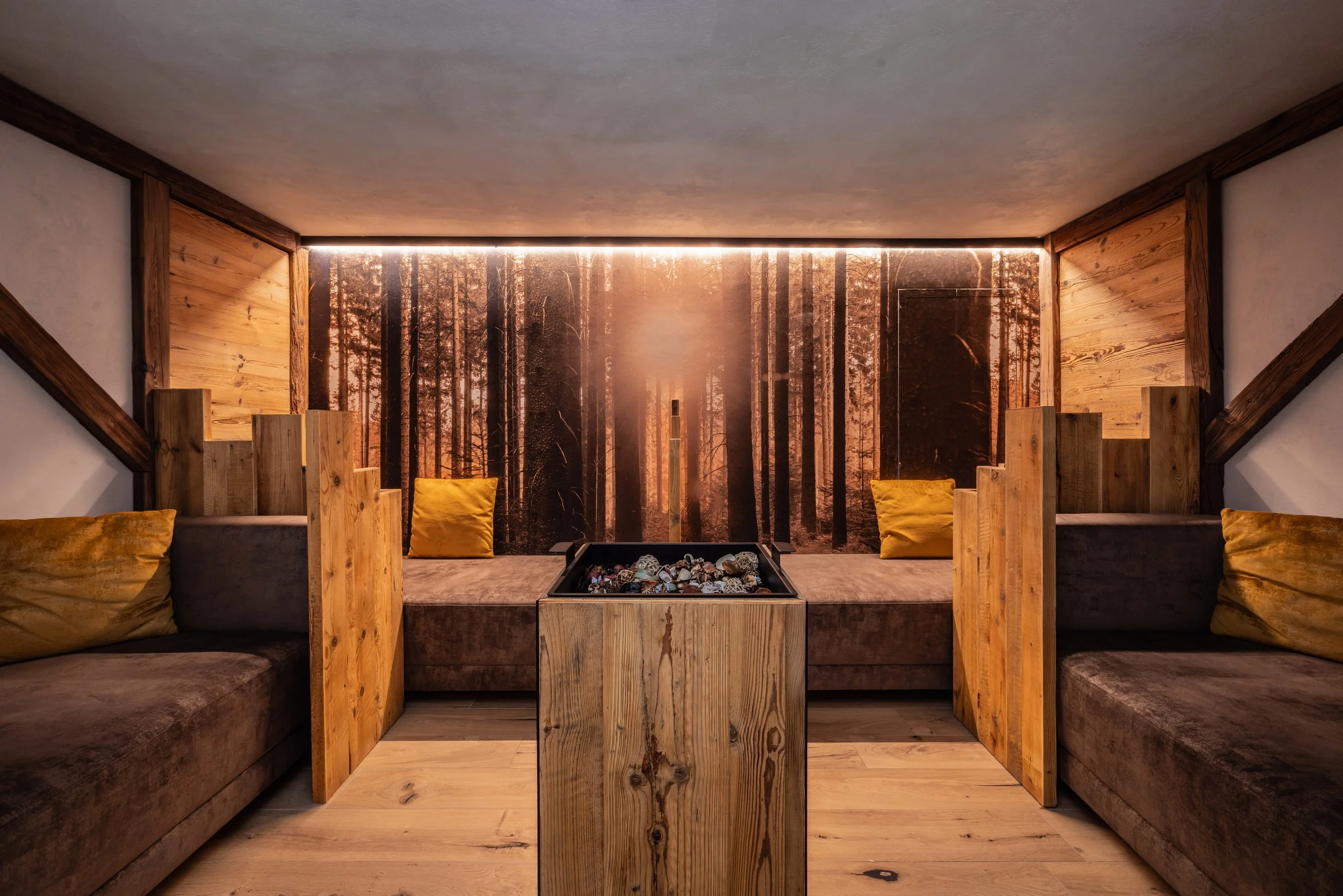 Interno di una stanza con mobili in legno e cuscini color senape, parete decorata con immagine di un bosco al tramonto.