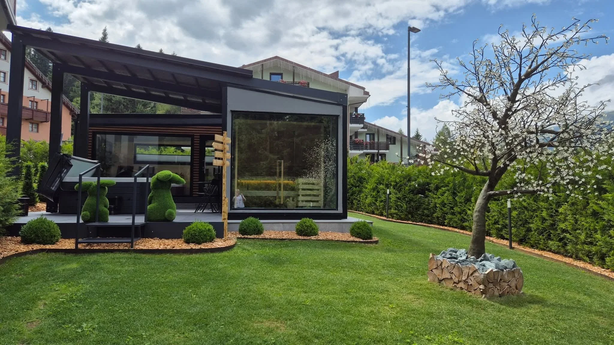 Giardino con gazebo moderno in vetro e legno, alberi fioriti, cespugli e un cortile ben curato, con case alpine sullo sfondo.