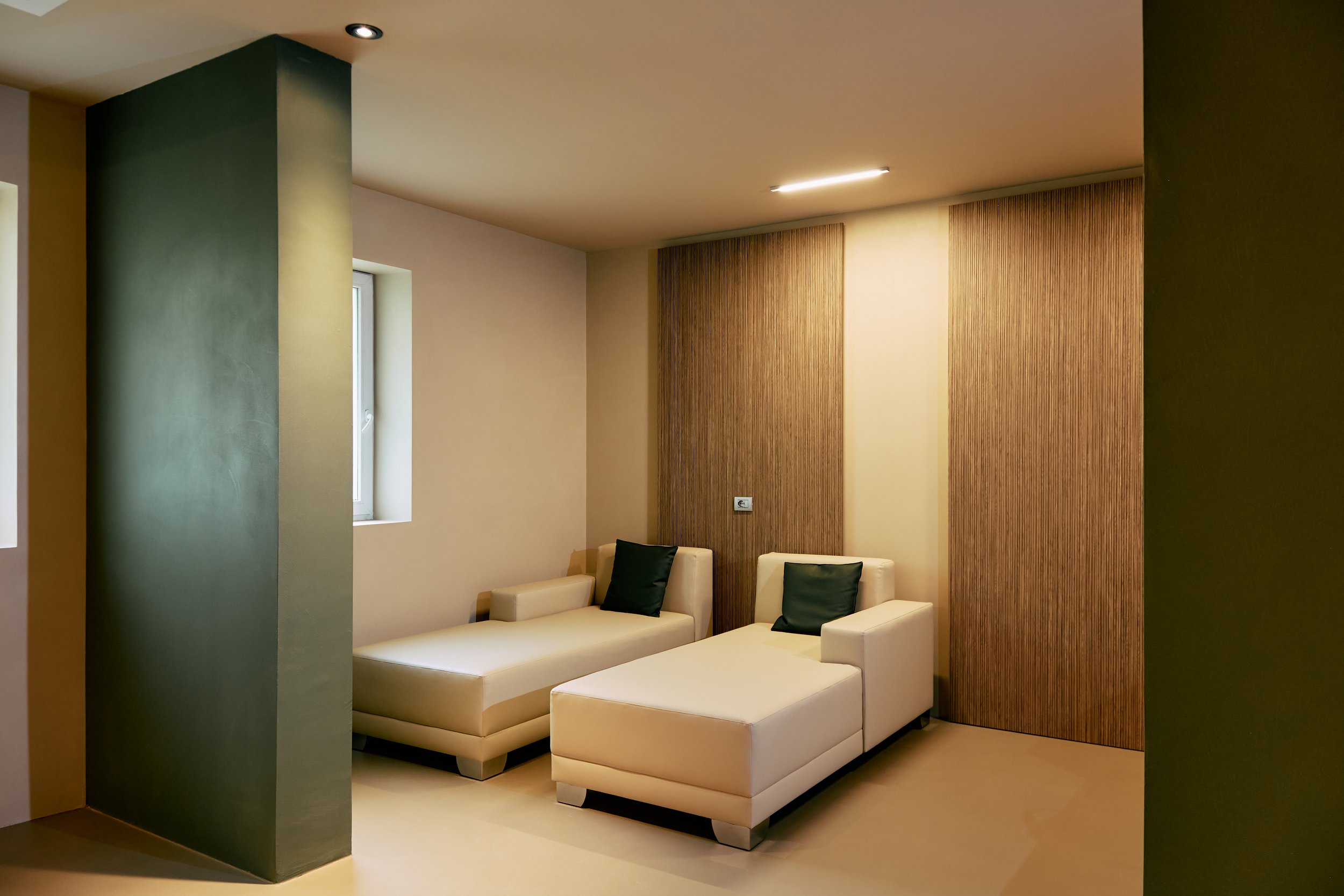 Interno con pareti beige e verde, pannelli di legno, divani bianchi con cuscini scuri, finestra.