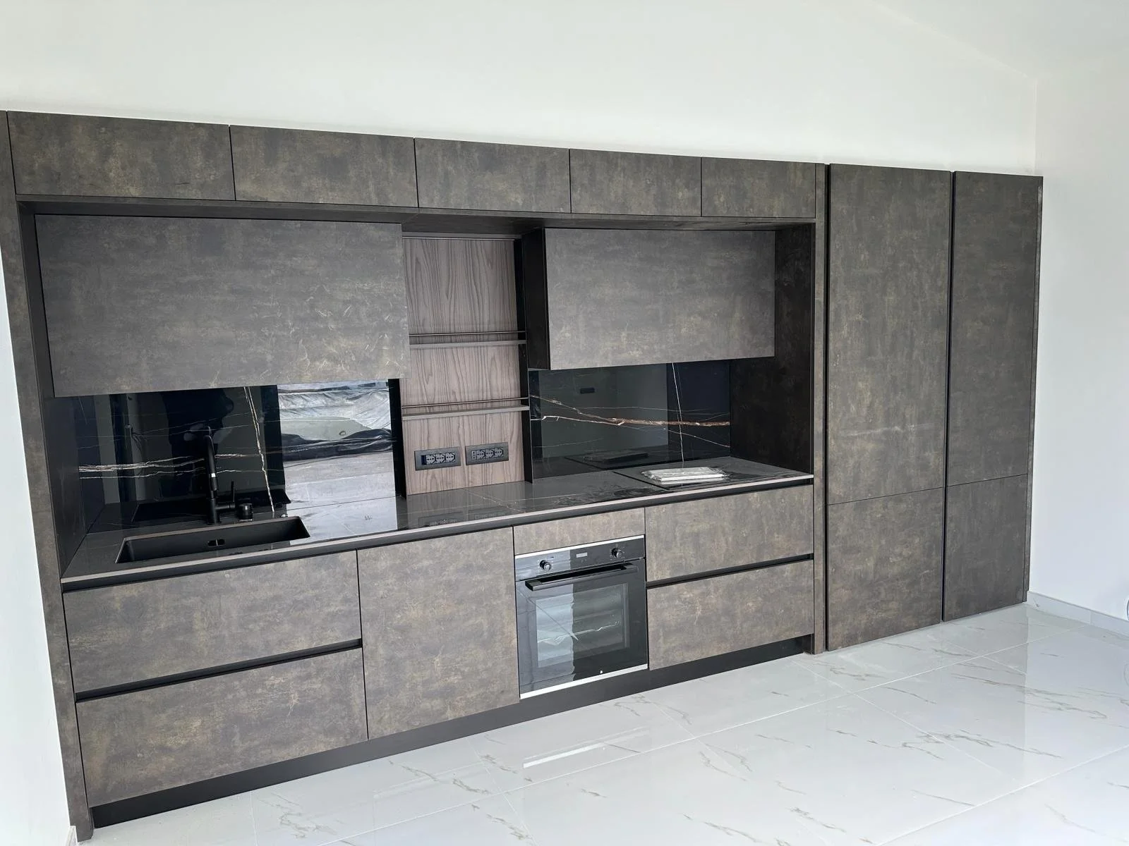 Cucina moderna con mobili in finitura scura e superfici in pietra, include un forno e piano cottura, con pareti bianche e pavimento in piastrelle chiare.