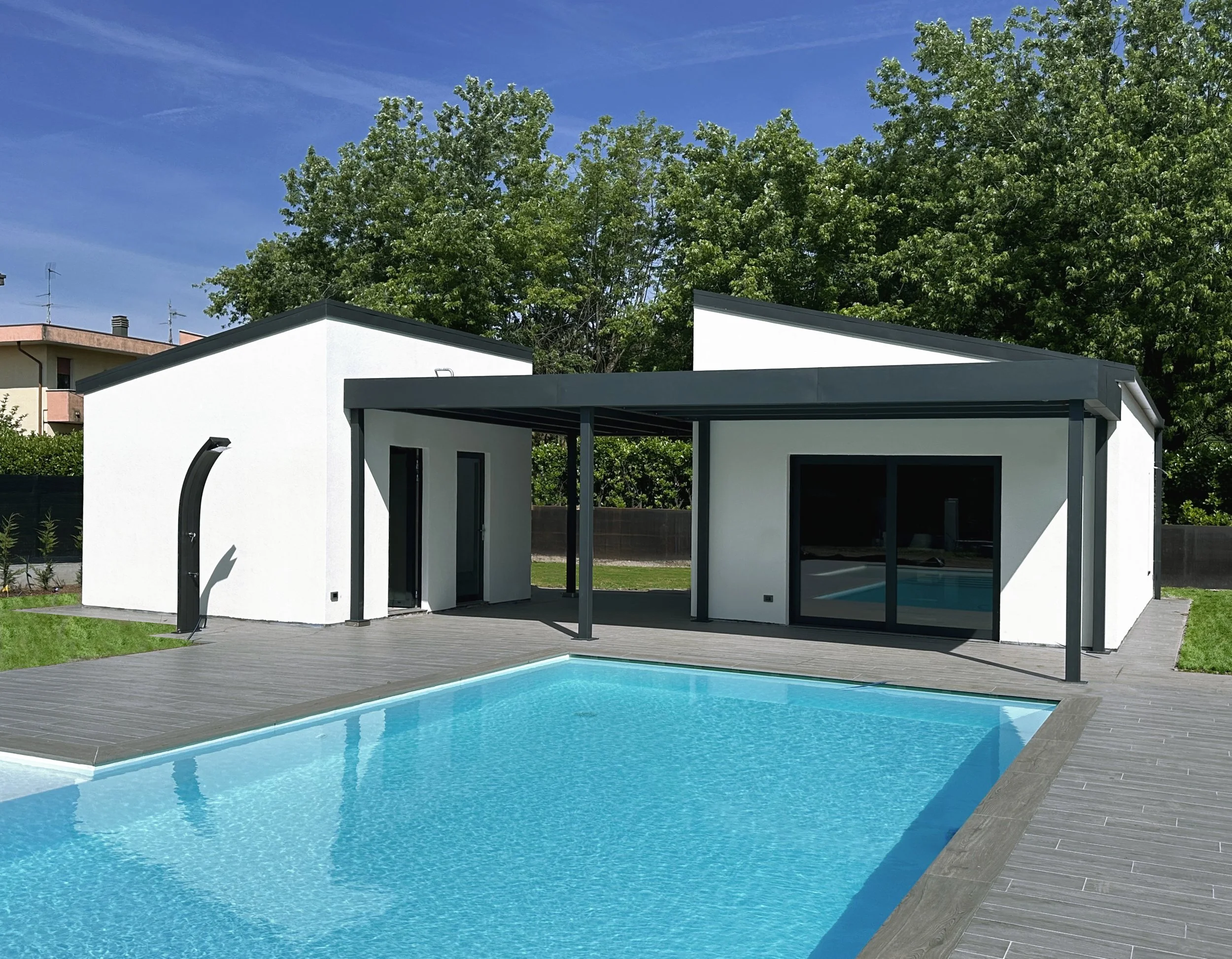 Piscina con bordo in piastrelle grigie, casa moderna con patio coperto, ampi vetri, e prato ben curato in un giardino con alberi verdi e cielo azzurro.