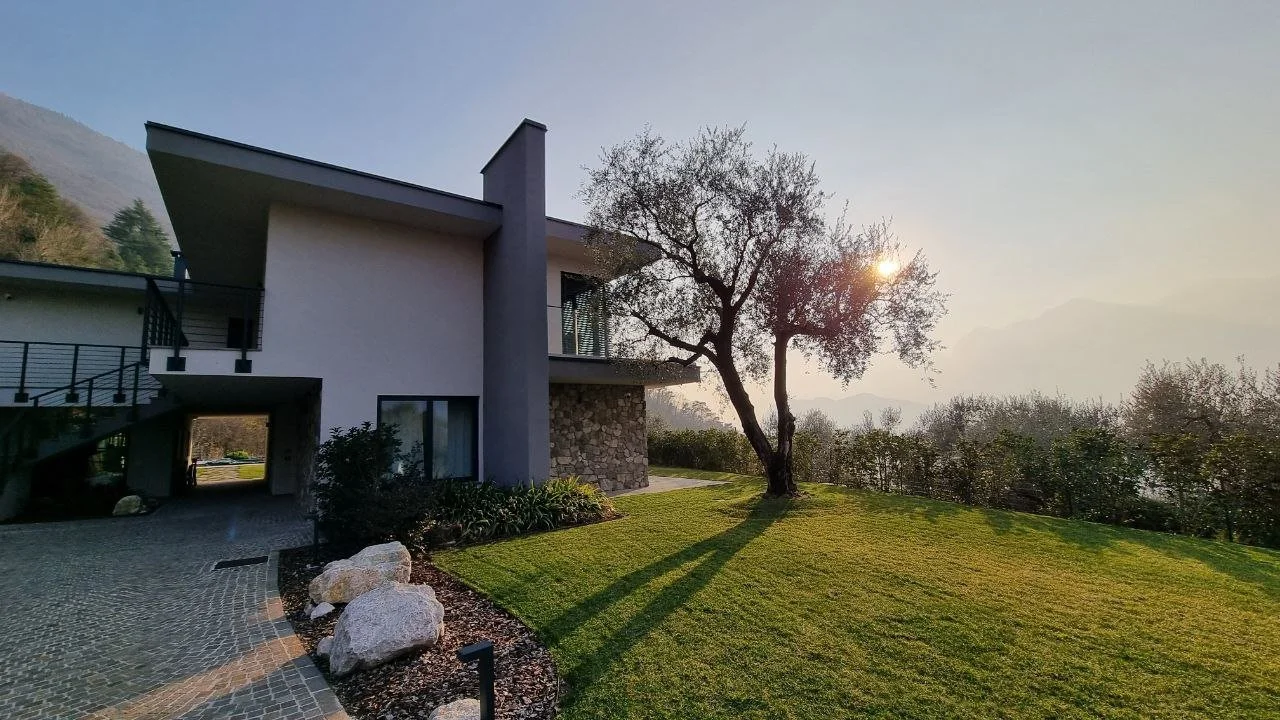 Casa moderna con giardino, albero e montagne sullo sfondo al tramonto.