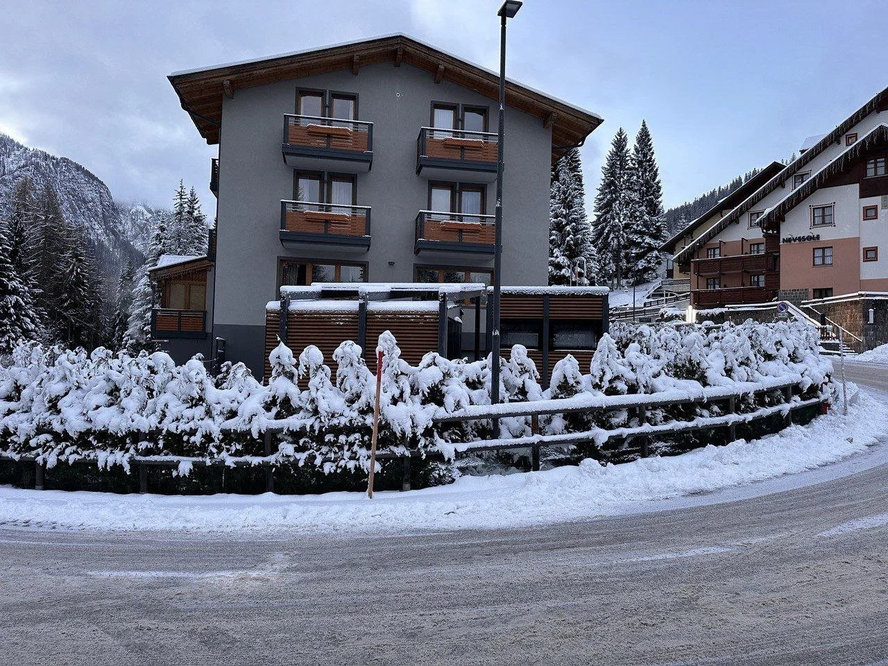 Edificio residenziale in montagna con neve, con balconi in legno, circondato da alberi e altri edifici, strada innevata in primo piano.