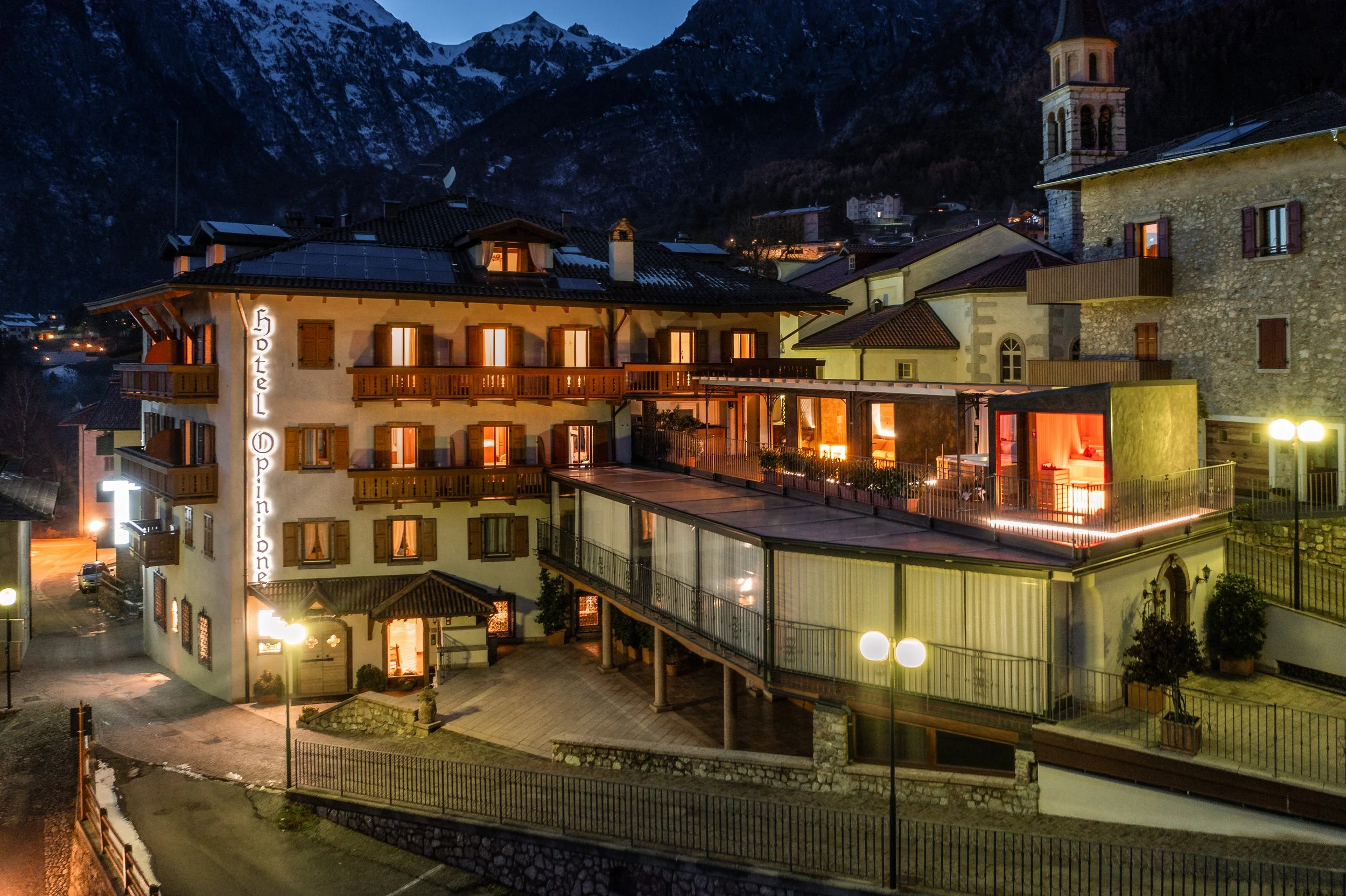 Paesaggio notturno di un villaggio alpino con edifici, montagne innevate sullo sfondo e luci calde emanate dalle stanze.