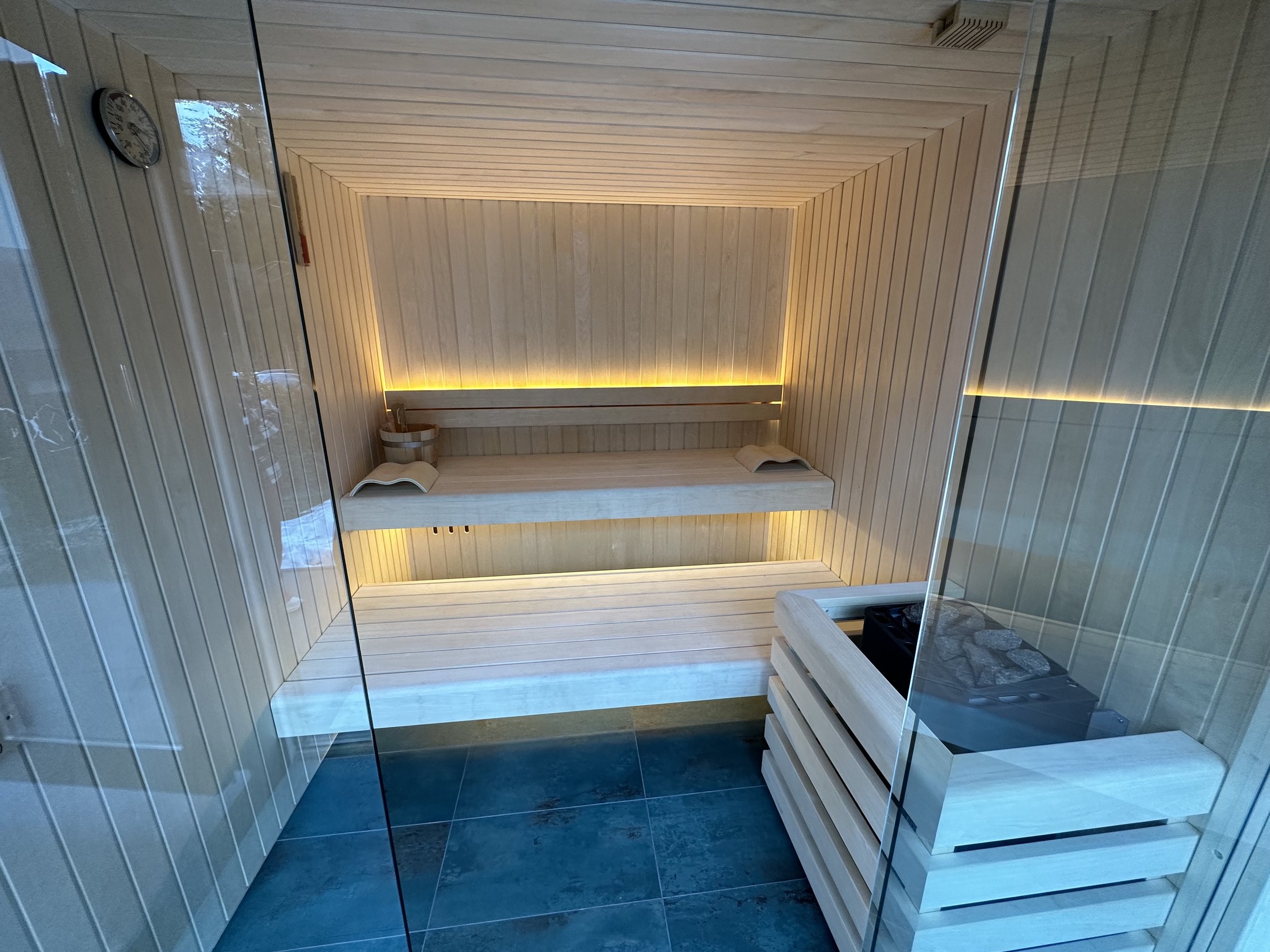 Interno di una sauna in legno con sedie e una luce calda lungo i bordi, visibile anche una brocca e un libro su una delle sedie.
