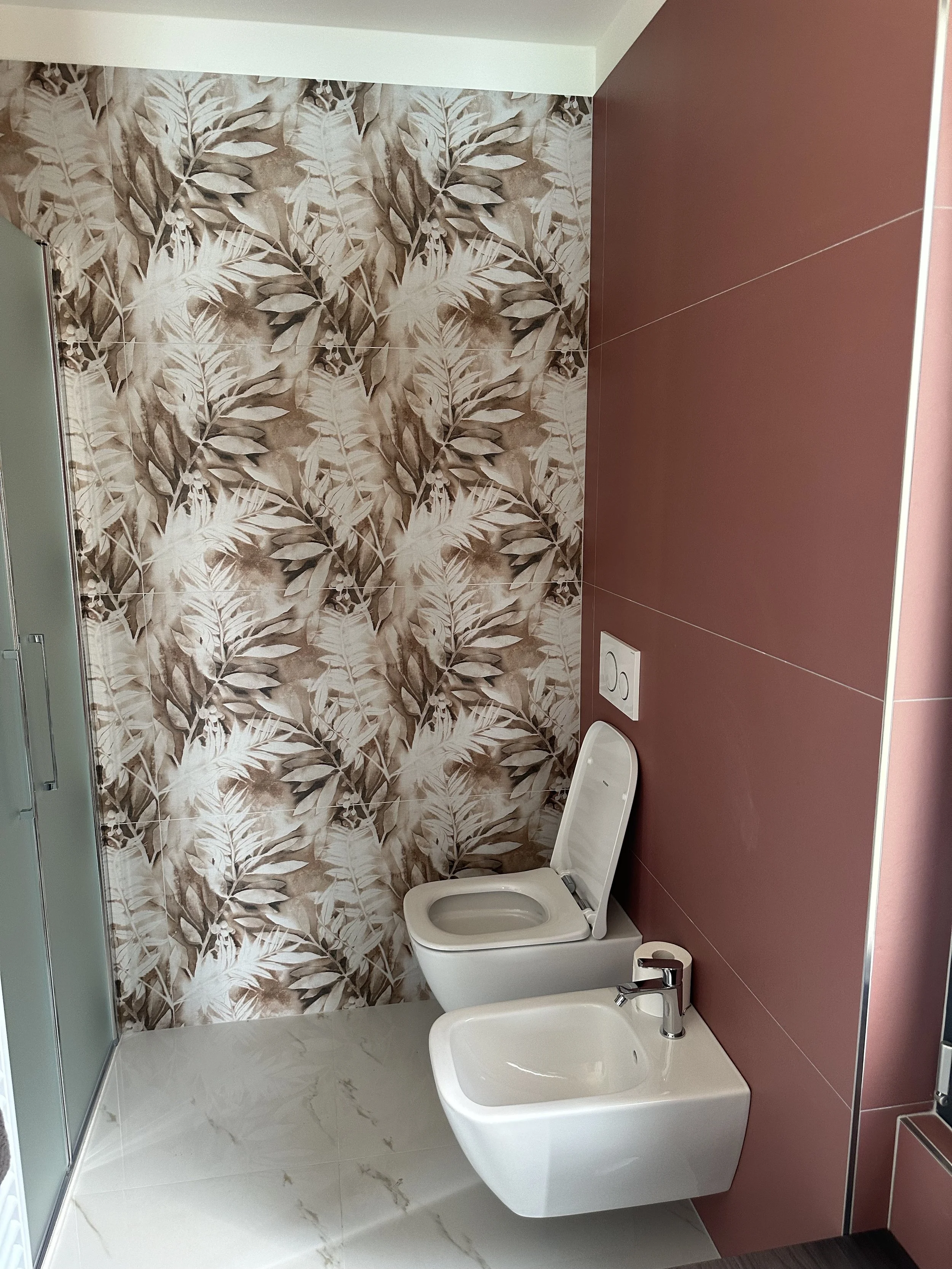 Bagno con parete decorata con carta da parati con motivi di foglie in toni marroni e beige, WC e bidet bianchi, lavabo con rubinetto, finestra sulla sinistra con vetro satinato, pavimento in piastrelle bianche con venature dorate.