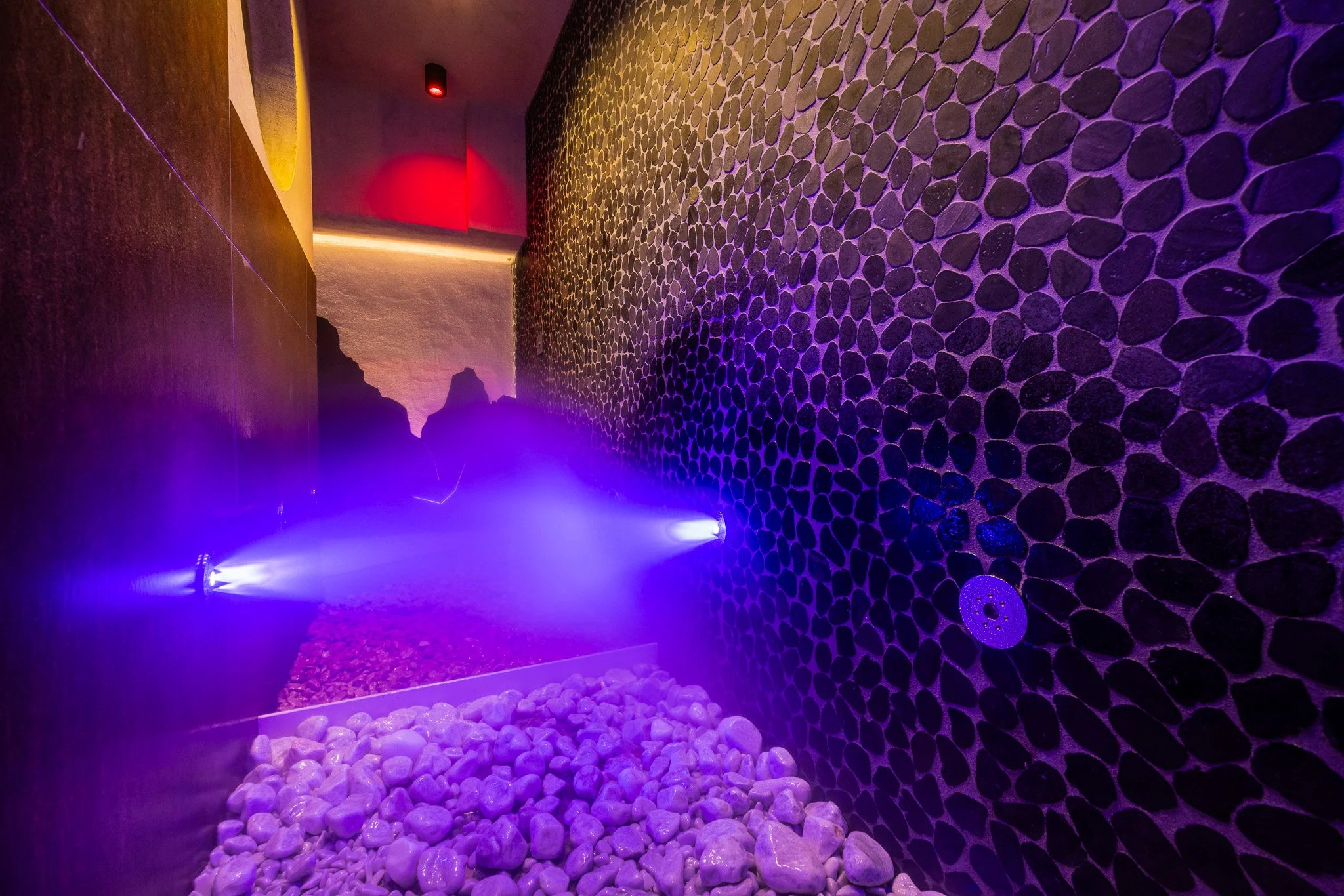 Un ambiente con pareti di pietre scure e luci colorate, illuminazione soffusa, pavimento con ciottoli lisci, atmosfera rilassante e moderna.