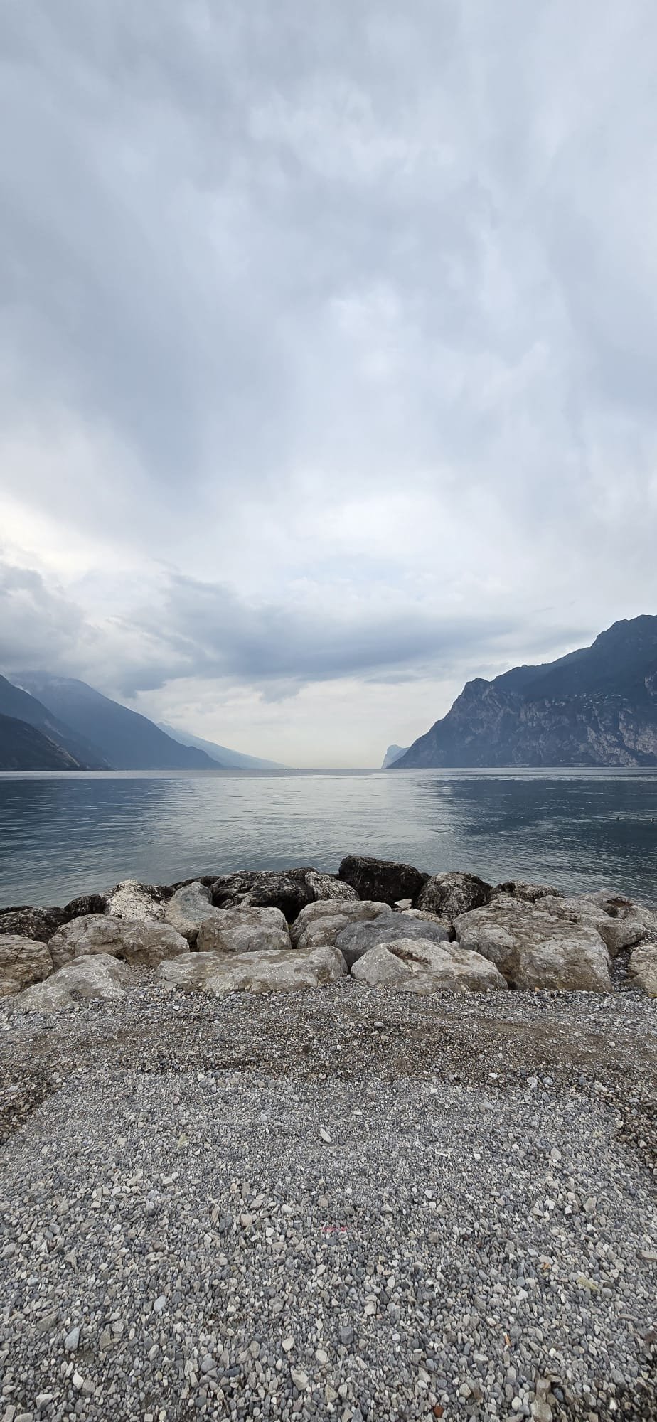 Lago circondato da montagne con cielo nuvoloso, vista dalla riva con grandi pietre e ghiaia in primo piano.