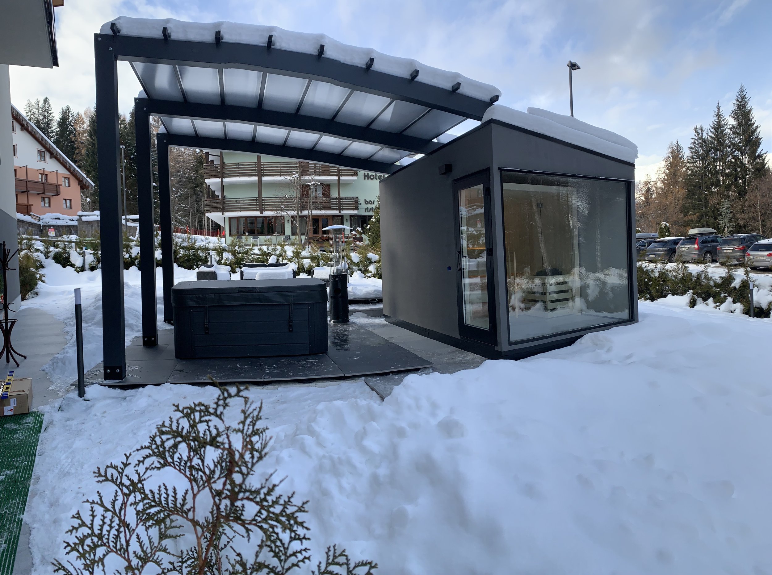 Struttura moderna in vetro e metallo in un'area innevata, con un edificio alberghiero sullo sfondo e un'area di parcheggio con automobili.