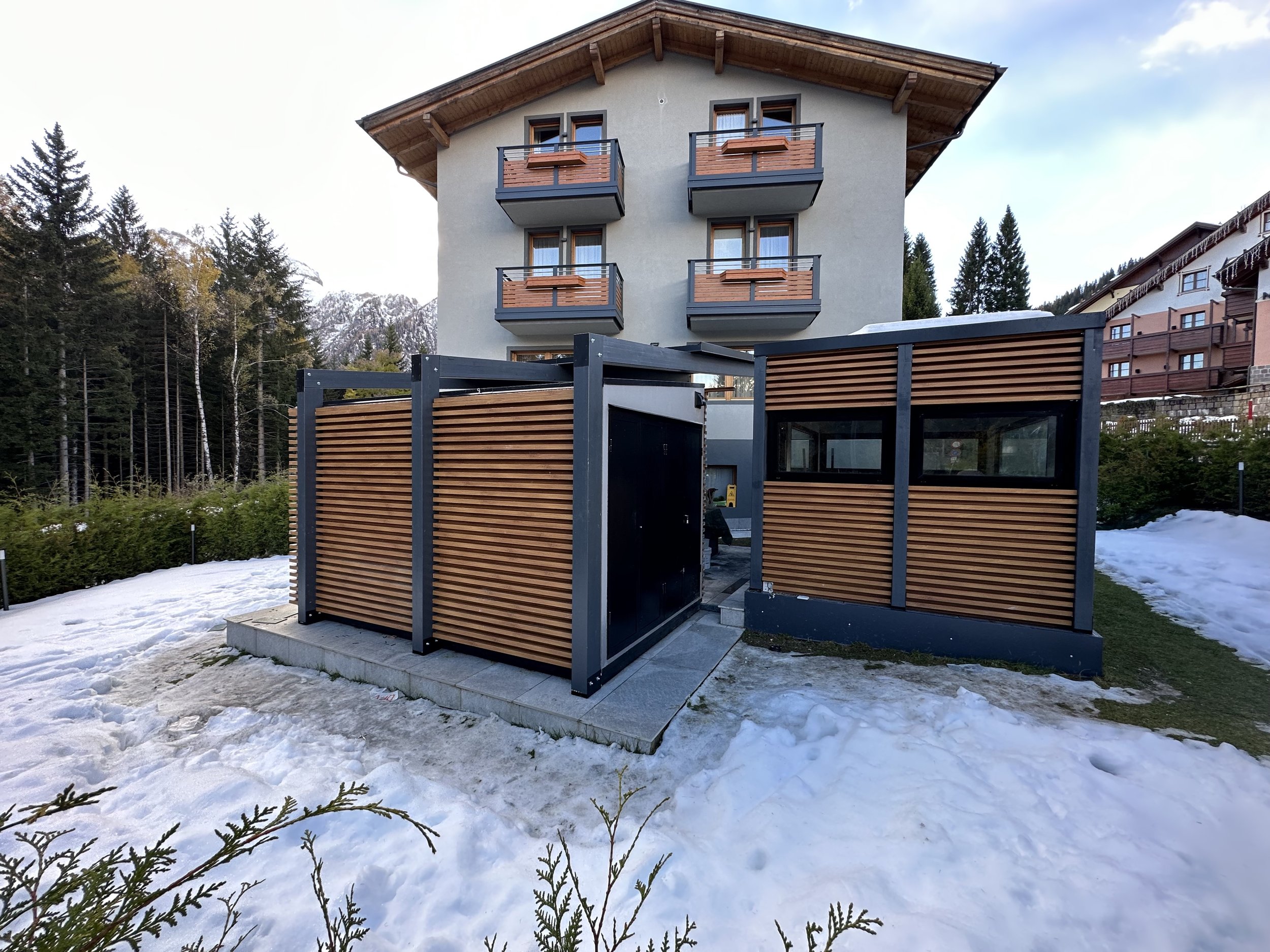 Edificio residenziale con balconi in legno e una recinzione in legno e metallo in primo piano, circondato da neve e alberi di conifere in montagna.