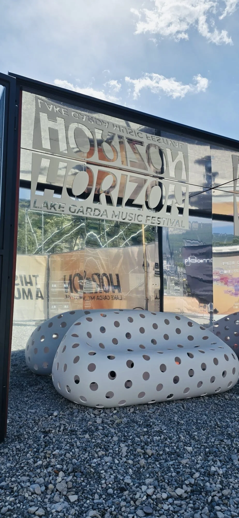 Segni di un festival musicale presso il Lago di Garda, con un'installazione artistica di sedili bianchi con fori neri e un cartello con il nome "Nozahon Horizon Lake Garda Music Festival".