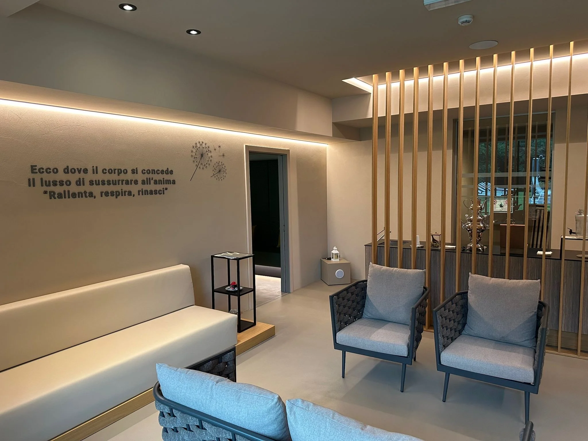 Interno di un ambiente moderno con sedie e divani in tessuto chiaro, parete decorata con scritta in italiano e decorazioni a forma di pigne, suddiviso da spinte di legno verticali.