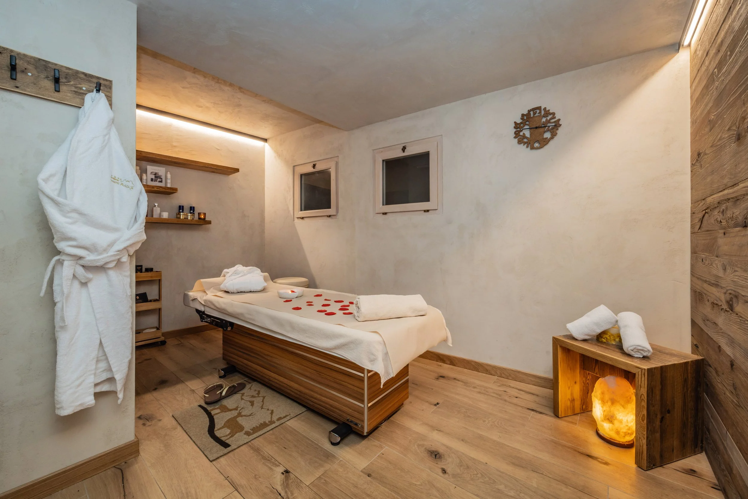 Stanza relax con lettino, decorata con ciotola di petali di rose e asciugamano arrotolato, lampada a sale, pareti di pietra e mobili in legno.