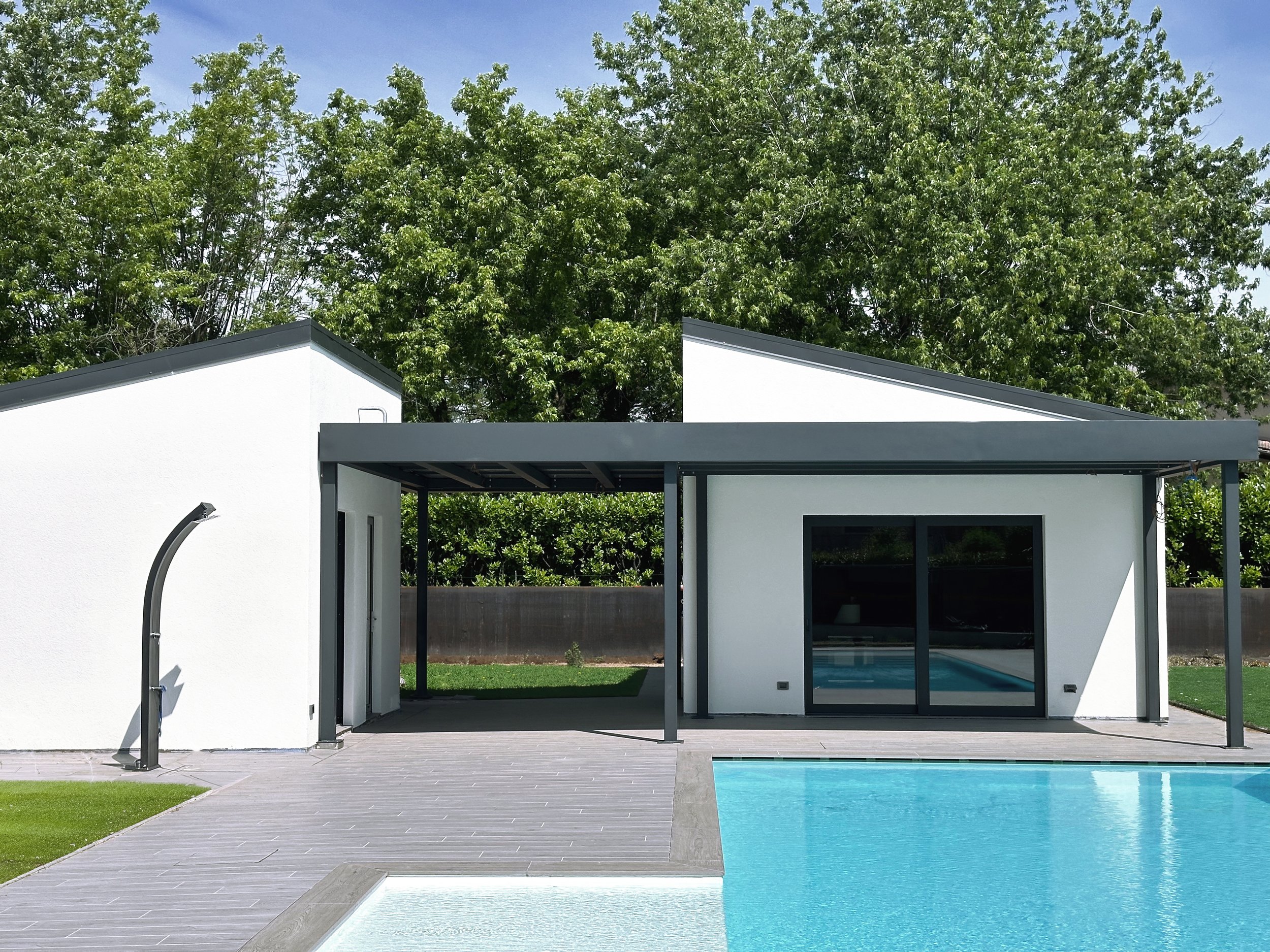 Vista di una casa moderna con patio e piscina, con grandi alberi verdi sullo sfondo e cielo sereno.
