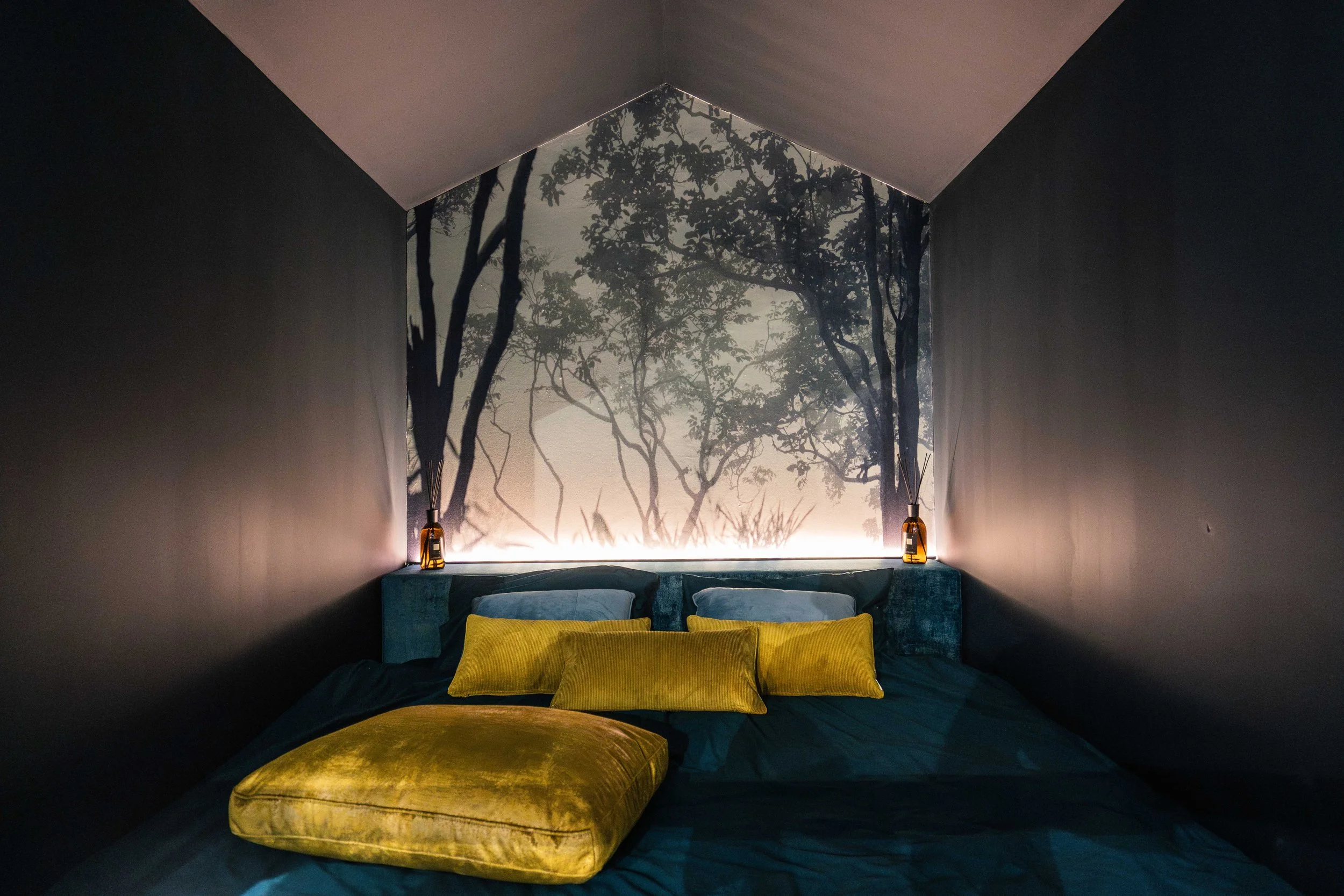 Letto con cuscini verdi in una stanza con pareti scure e una grande decorazione murale con un albero stilizzato dietro. Sul comodino ci sono due lampade di vetro e un soffice piumone blu scuro.