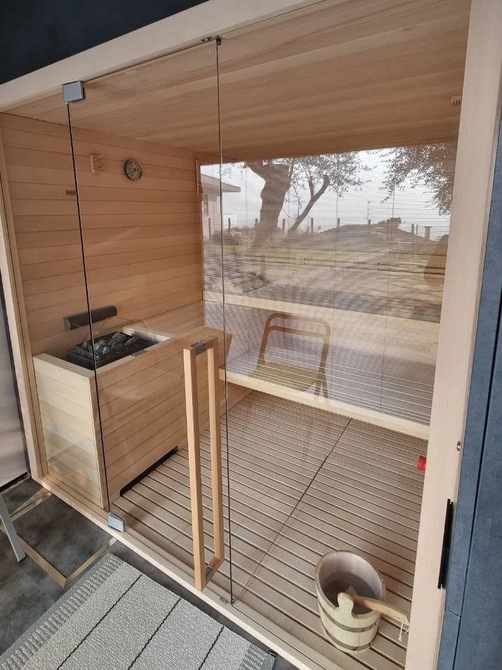 Sauna in legno con pareti di vetro divisori, vista sulla natura esterna con alberi e terreno, presente un secchio con paletta e un orologio sulla parete.