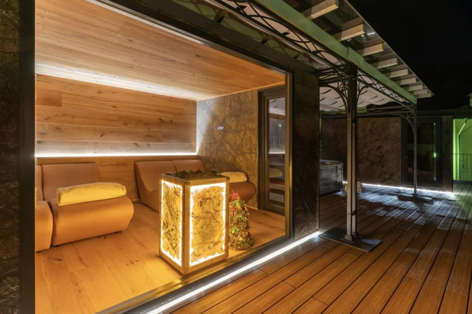 Veranda moderna con pareti e soffitto in legno, illuminazione LED e mobili in stile contemporaneo, con un divano, una lampada decorativa illuminata, e un'area esterna in legno con luci soffuse.