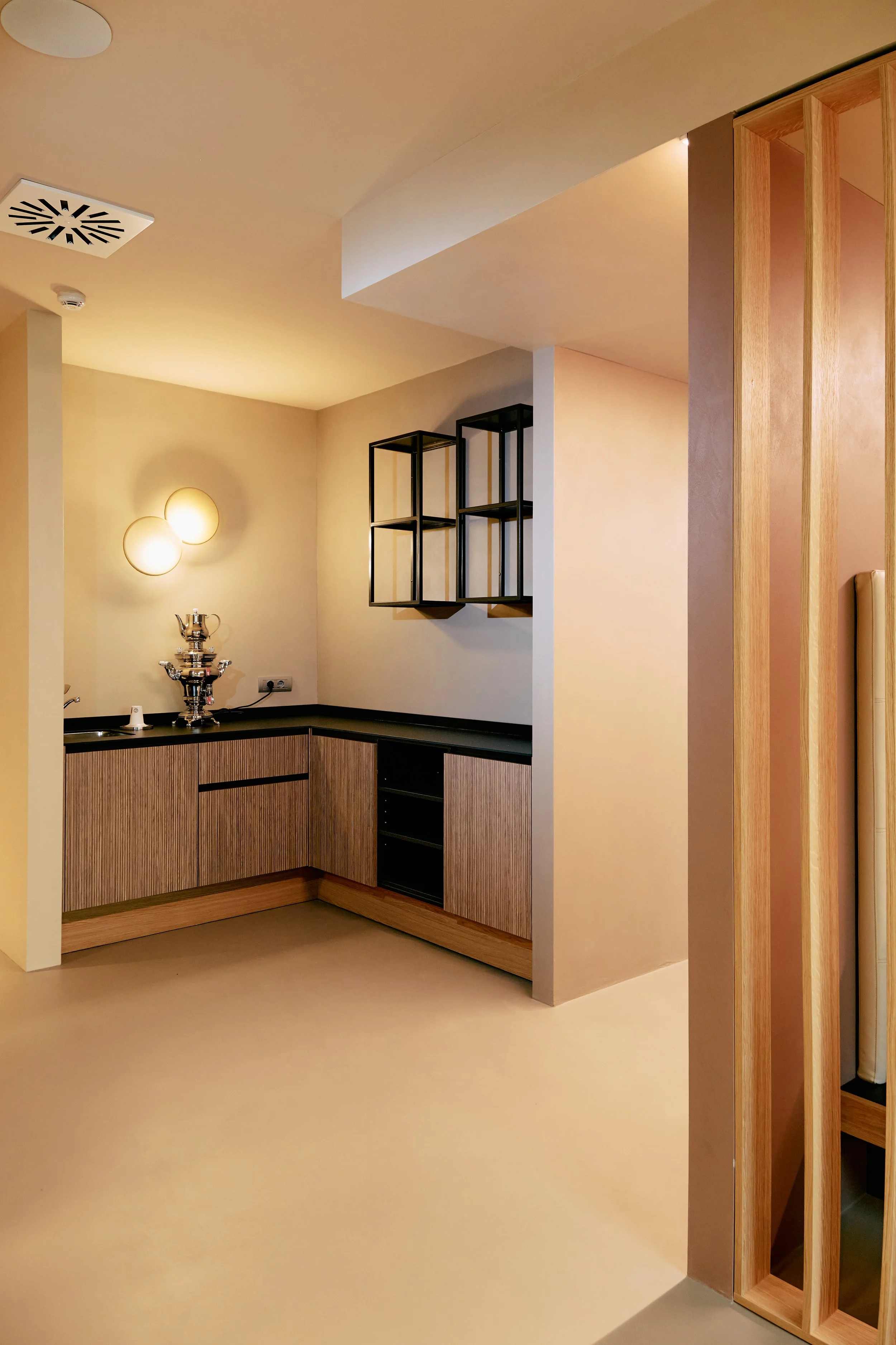 Interno di una cucina moderna con pareti beige, un mobile sospeso con mensole in metallo nero e un bancone in legno chiaro. Lampade a parete decorano la parete sinistra e sul bancone è presente una teiera in metallo.
