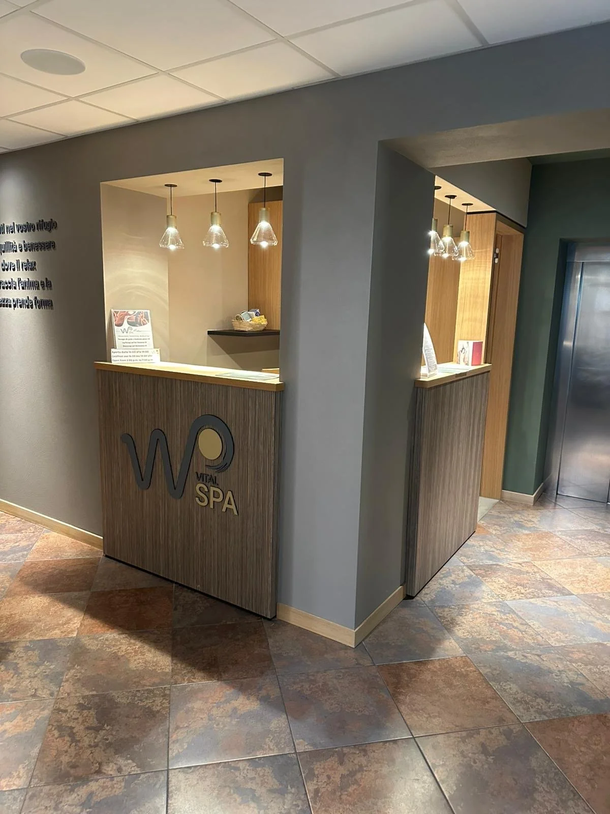 Reception del centro benessere WP Vital SPA con pareti di colore grigio, reception in legno con logo visibile e lampade decorative sopra. Pavimento di piastrelle in tonalità calde.