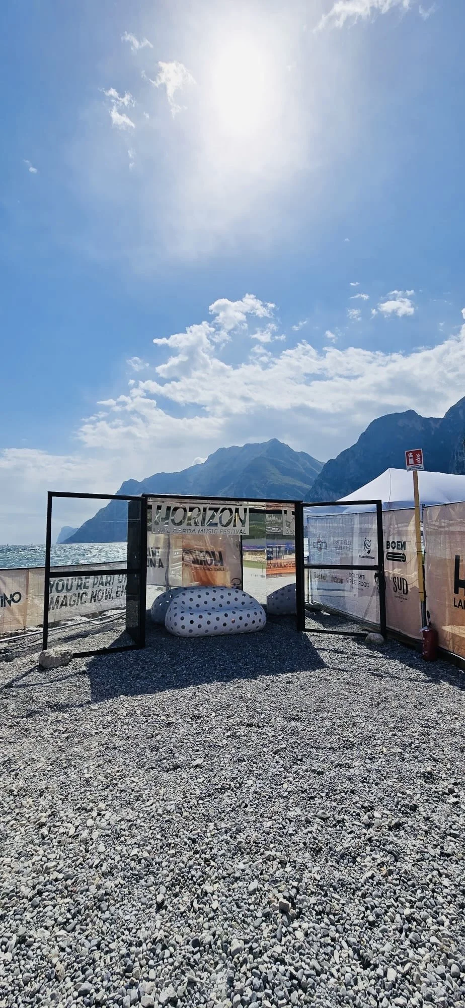 Spiaggia con fondale montuoso, cielo soleggiato, area delimitata da barriere con banner promozionali e sedili gonfiabili a pois.