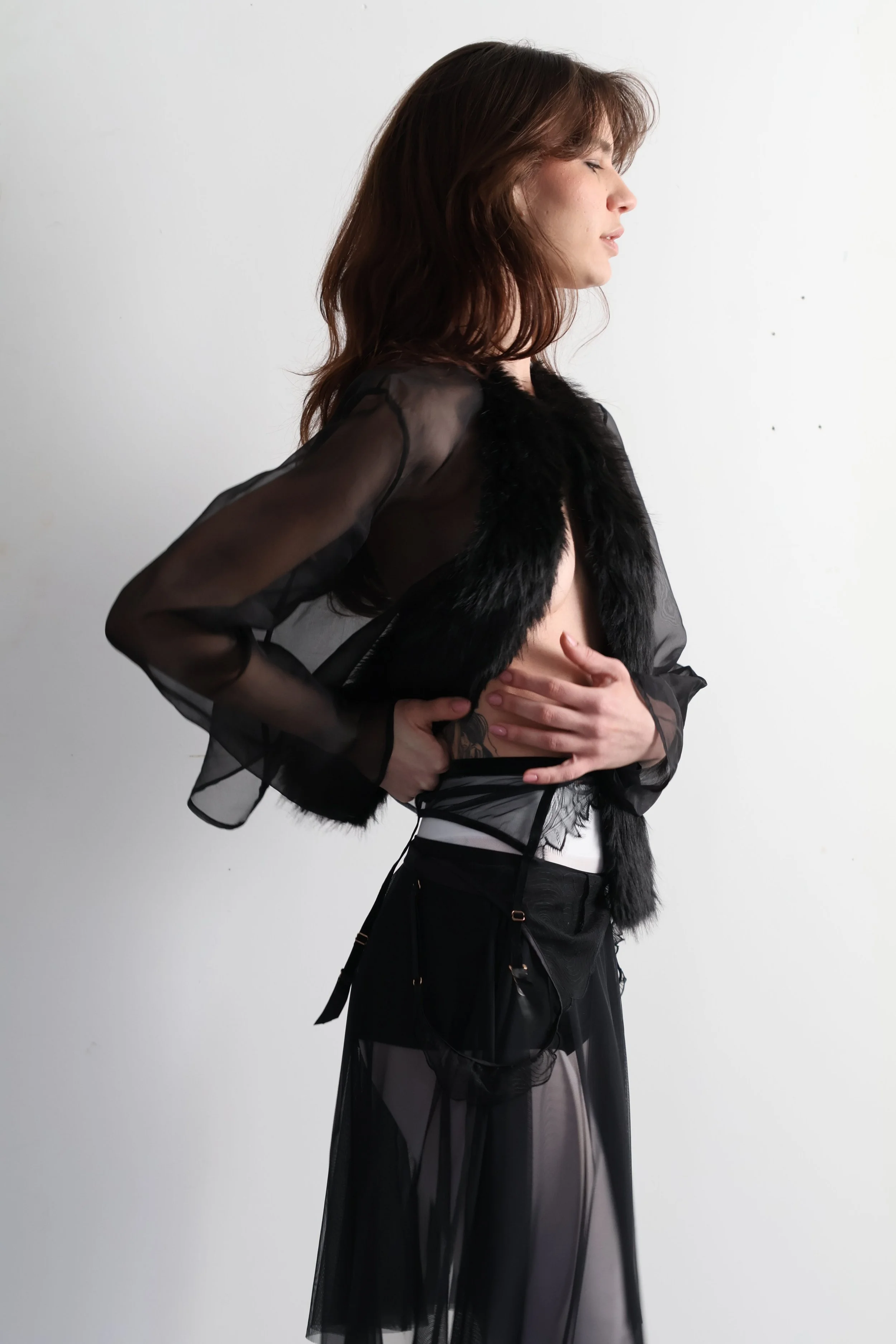Mulher de perfil com cabelo castanho, usando roupa preta de pano transparente e peles, posicionada contra parede branca. Parte de editorial de moda sobre intimidade e transparência.