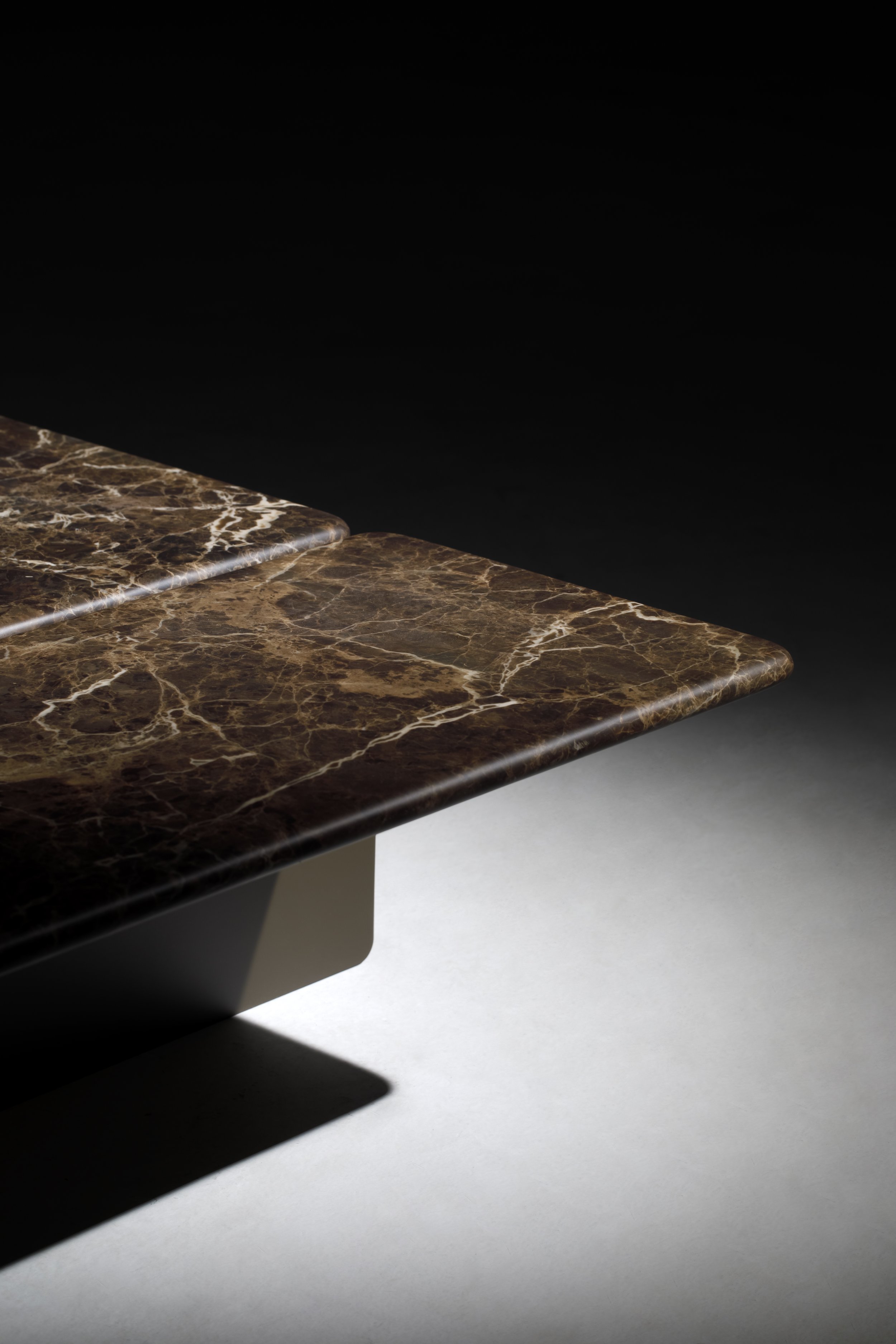 Mesa de centro de mármore com veios brancos e pretos, com o canto de uma mesa visível sobre uma superfície branca, e uma sombra projetada por ela.