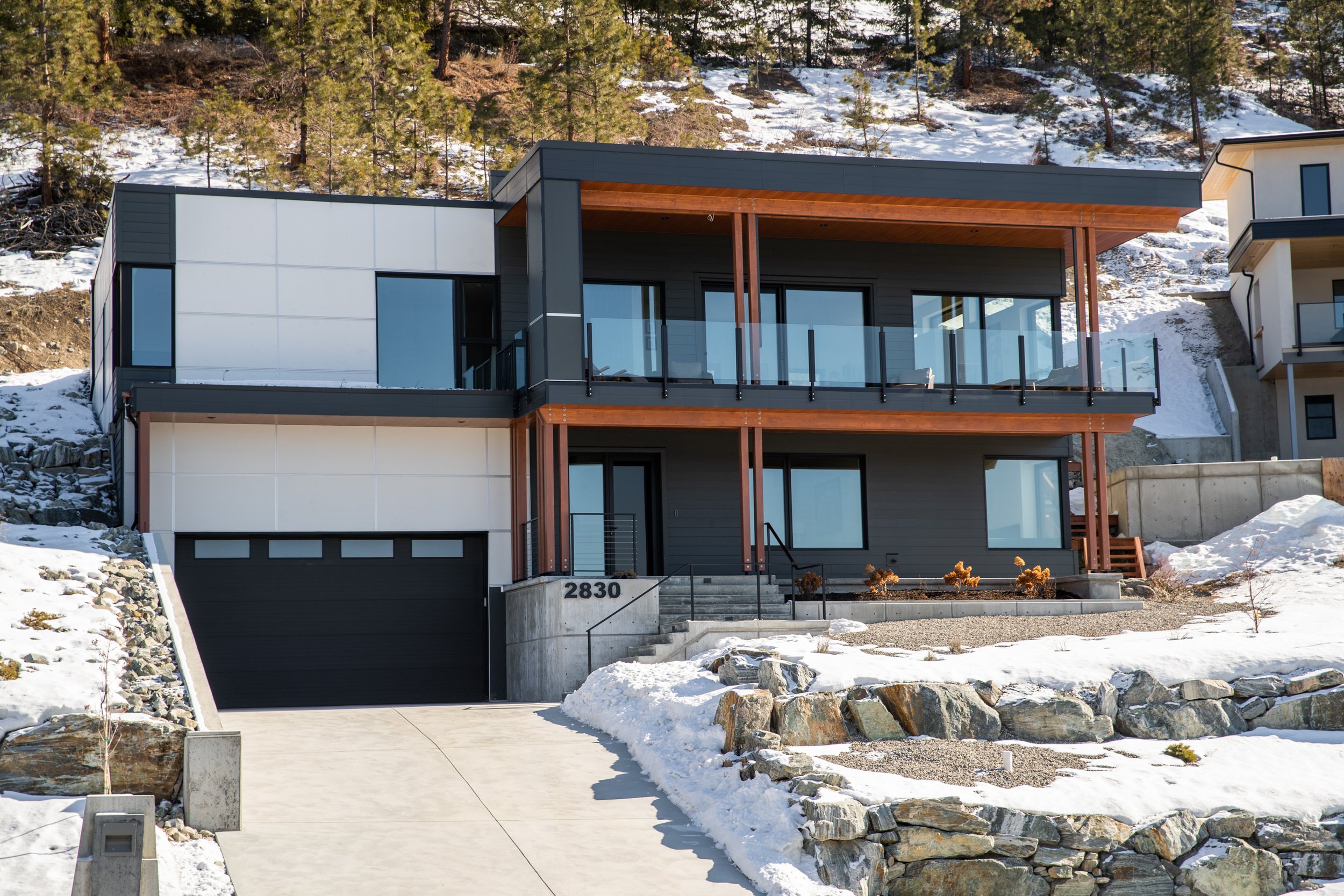 Custom built house on the Naramata Bench with large glass balcony overlooking snowy landscape and pine trees, located on a hillside with driveway and concrete steps.