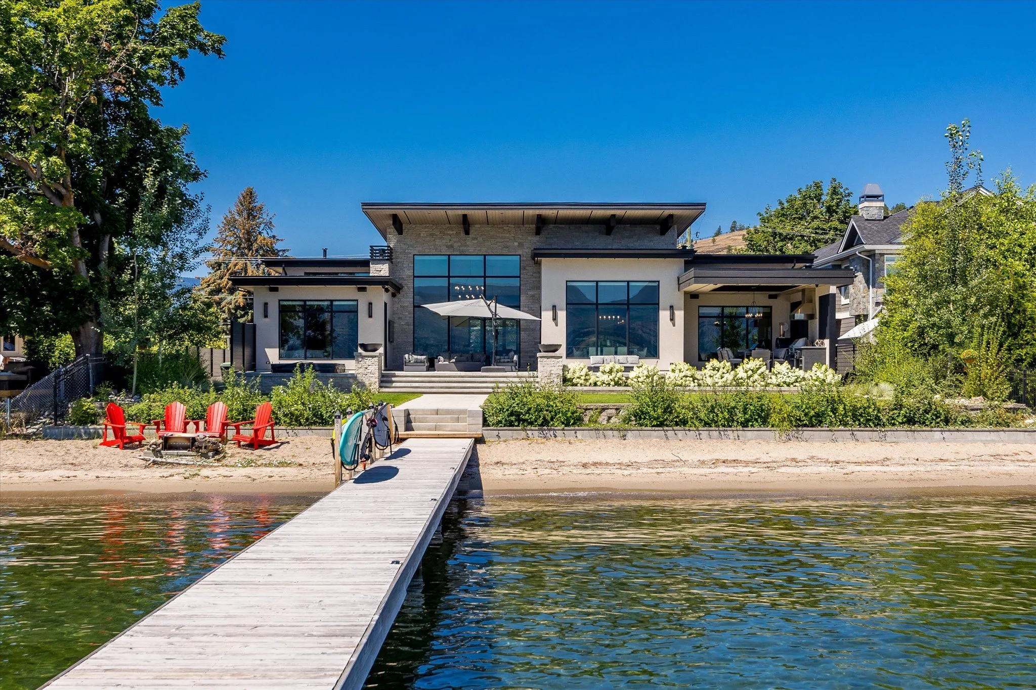 An award winning home on Okanagan Lake's Pritchard Drive with large windows and a backyard patio, situated next to a body of water with a wooden dock extending into the water, surrounded by trees and greenery.