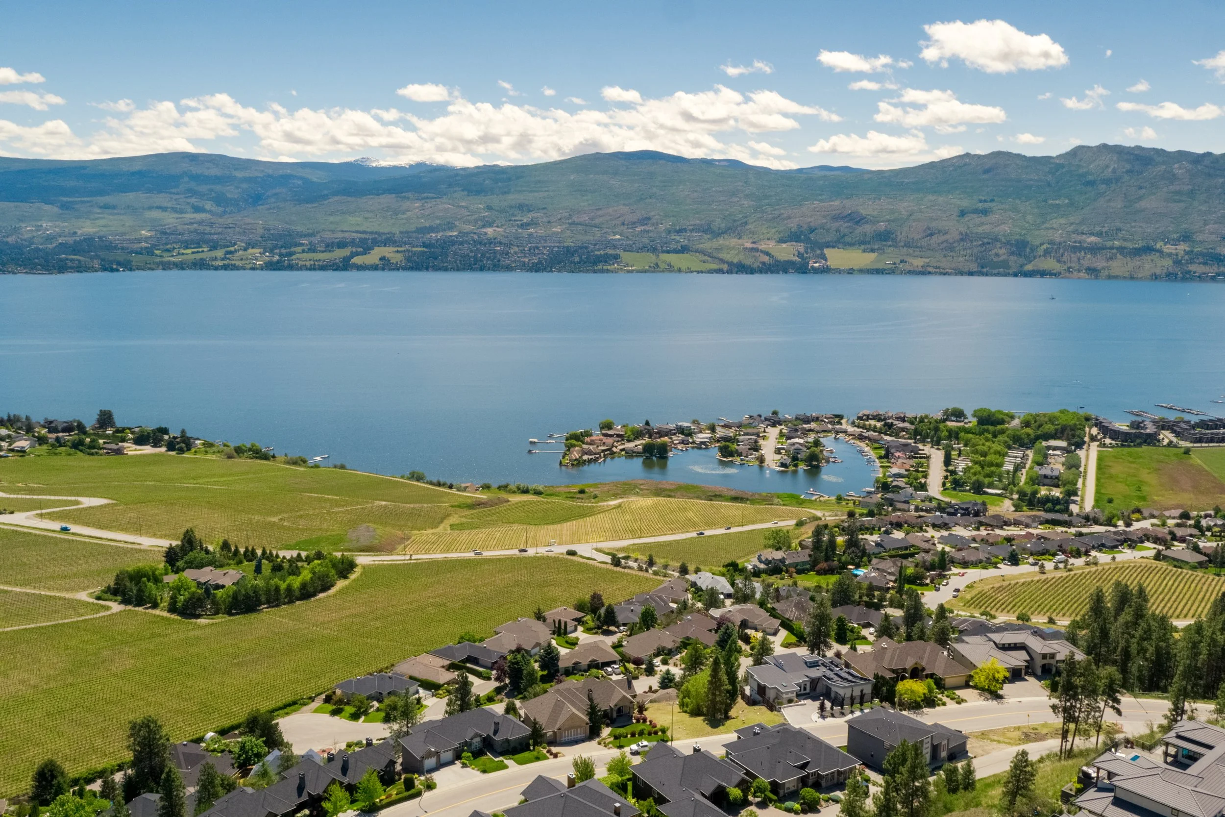 Green Bay aerial view of a residential neighborhood with houses, a lake, green fields, and mountains in the background under a partly cloudy sky.