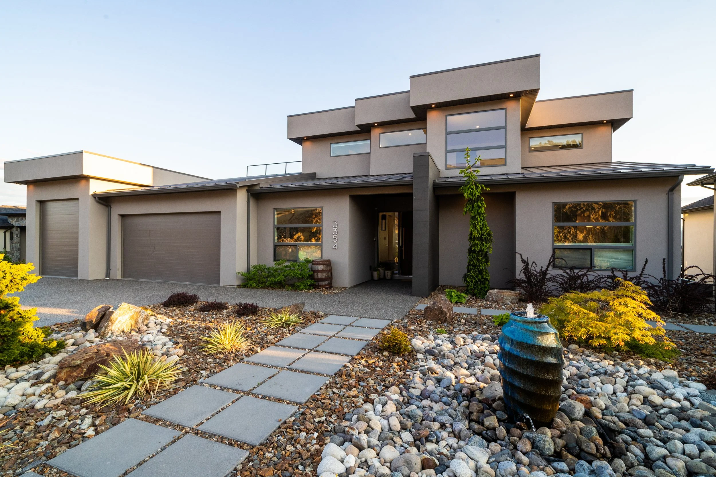 Modern two-story house with a dark gray front door, garage, large windows, and landscaped front yard with a stone pathway, rocks, plants, and a blue water fountain.