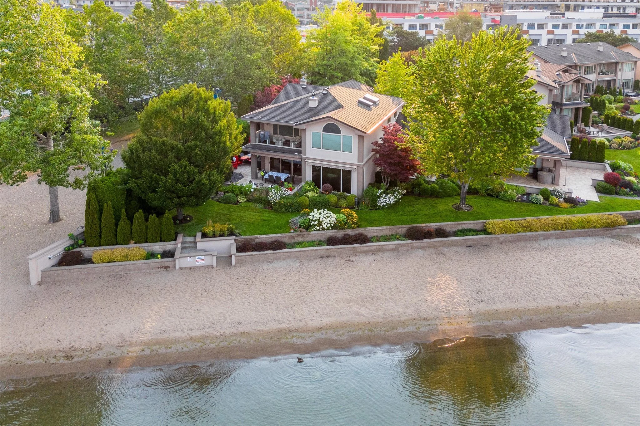 An aerial view of a townhome on Okanagan Lake surrounded by tall green trees and a well-landscaped garden, with a waterway at the bottom of the image.