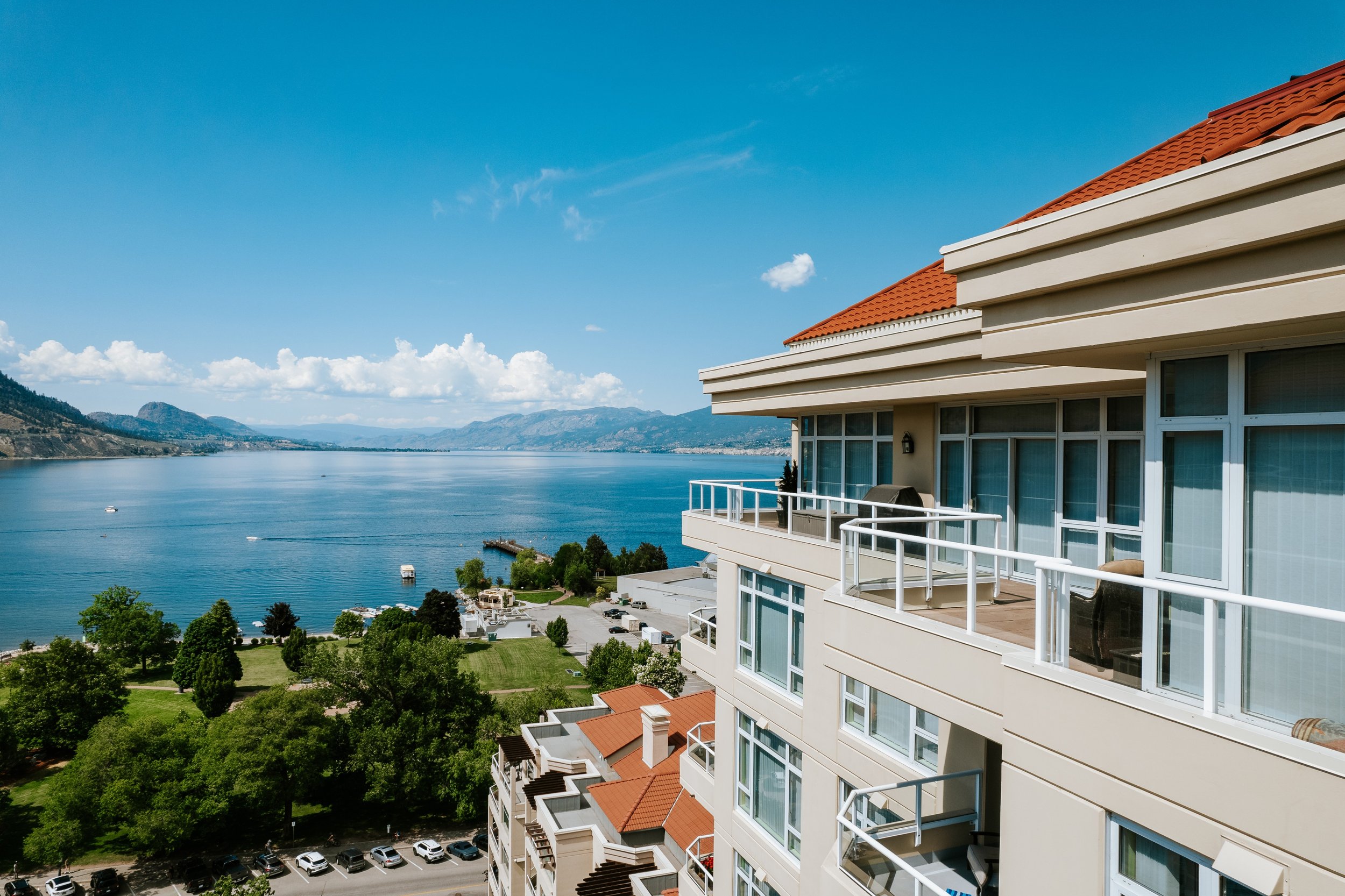 A view from a balcony of a modern condo in Penticton British Columbia with a view of Okanagan Lake and mountains in the background. The building has white walls, large glass windows, and red-tiled roofs.