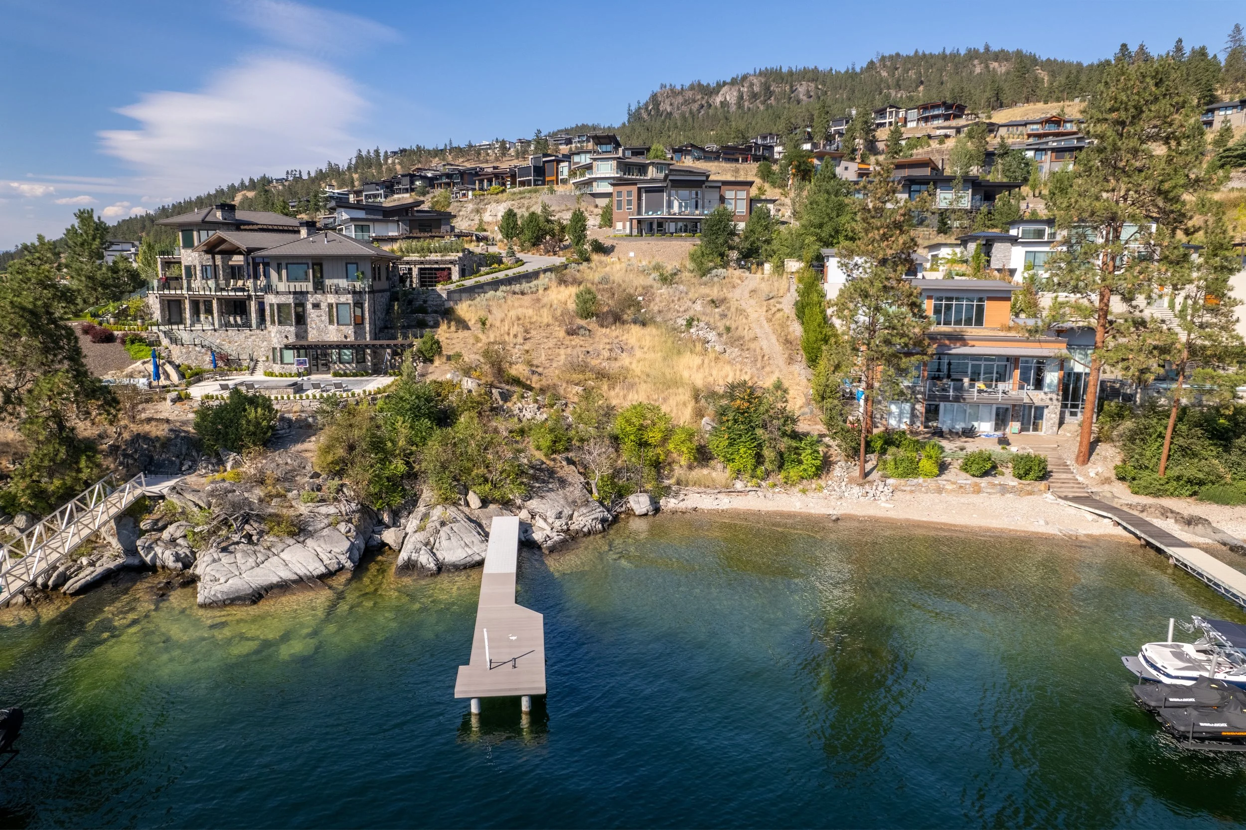 Aerial view of a building lot in Lake Country on Okanagan Lake with a dock extending into the water and surrounding trees, under a blue sky.