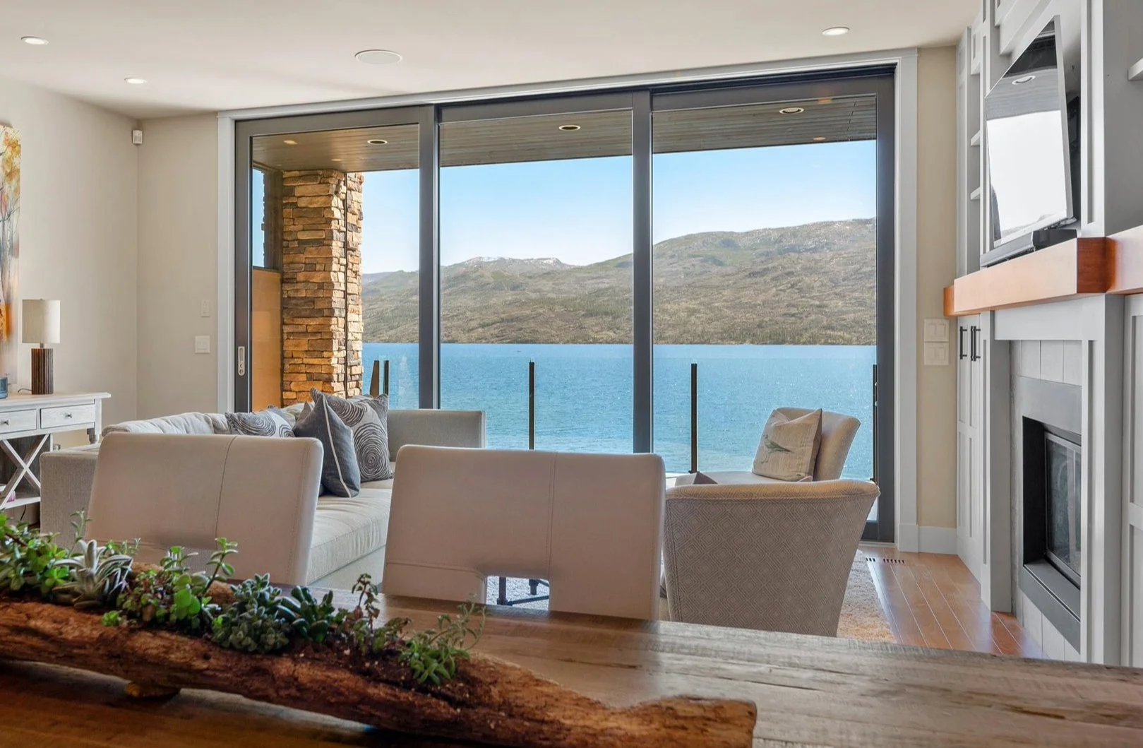 Peachland Townhome on Okanagan Lake with a living room with large glass sliding doors showing a lake and distant mountains outside. Inside, there are white couches, an armchair, a fireplace, and a TV. 
