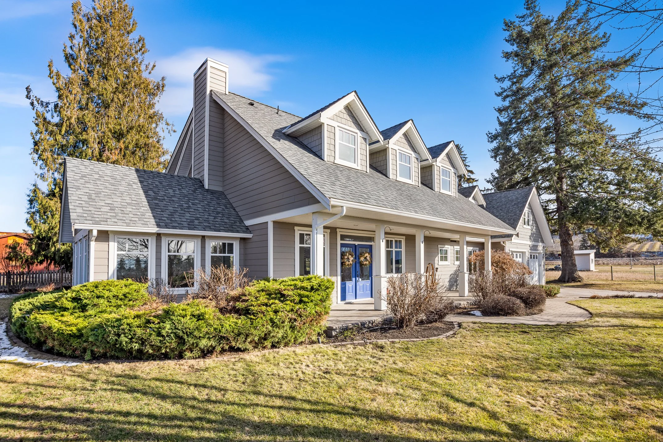 A two-story house family home in South East Kelowna with a gabled roof surrounded by a front yard with green shrubs and a lawn. There are trees behind the house and a bright blue sky overhead.