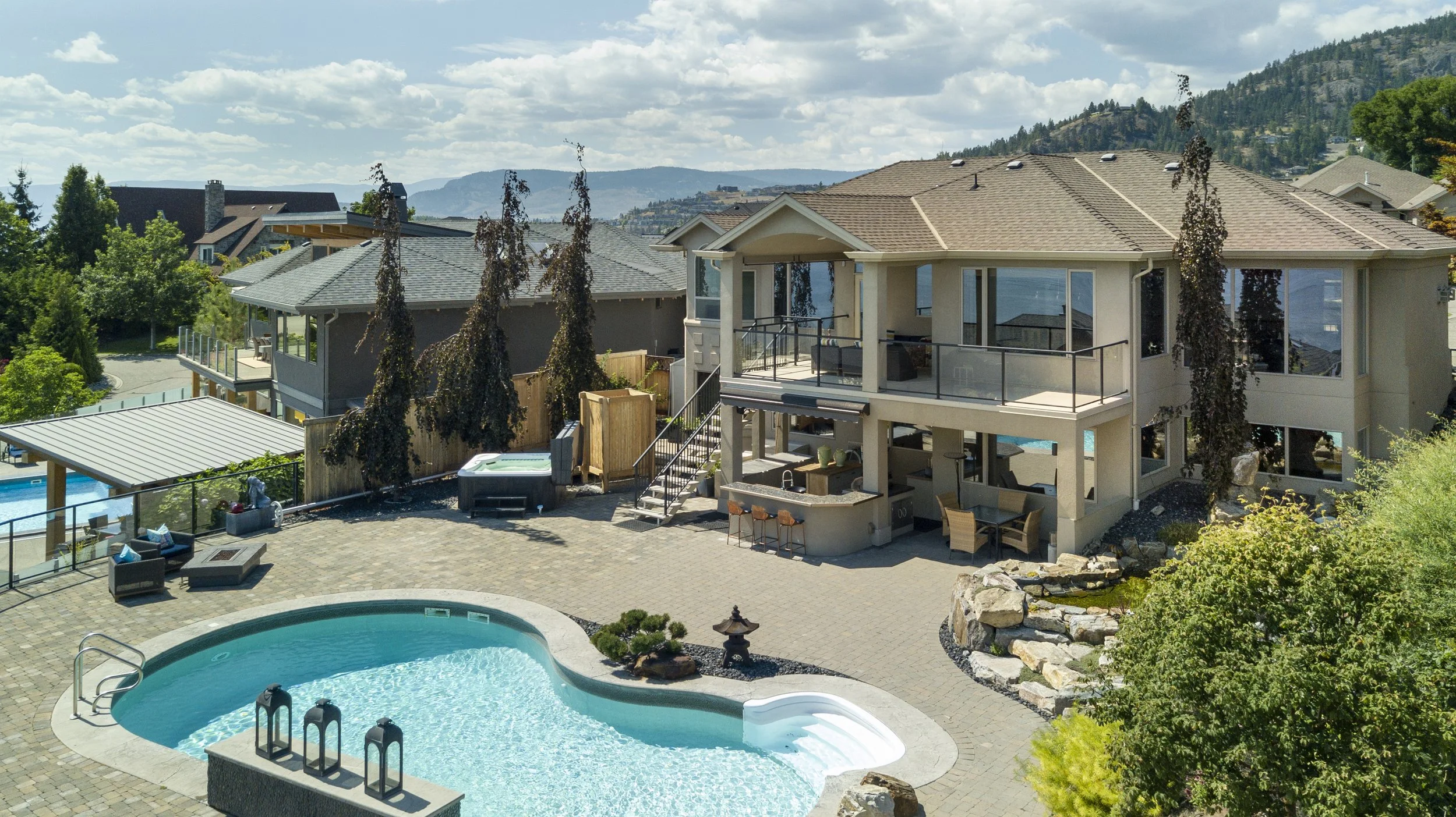 A West Kelowna backyard with a in-ground swimming pool, hot tub, outdoor seating, a hot tub, and a modern two-story house with a deck and large windows, surrounded by trees and mountains.