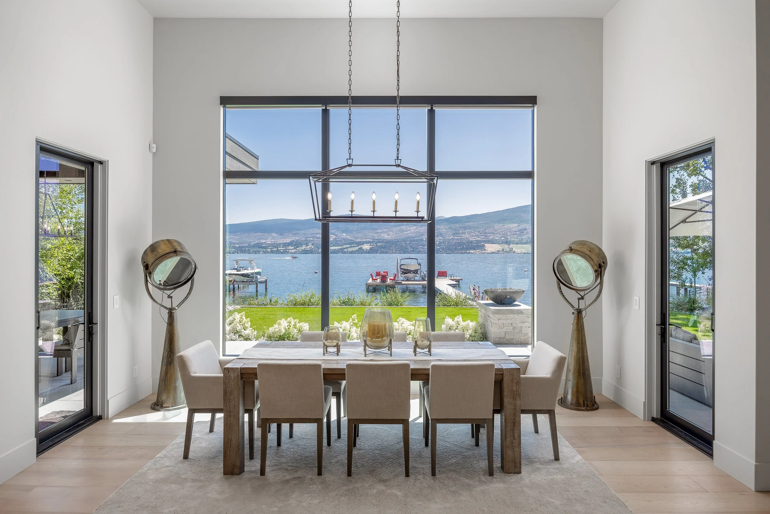 Modern dining room with large floor-to-ceiling windows overlooking a lake and mountains, featuring a wooden dining table with six light-colored chairs, a chandelier, and decorative lamps.