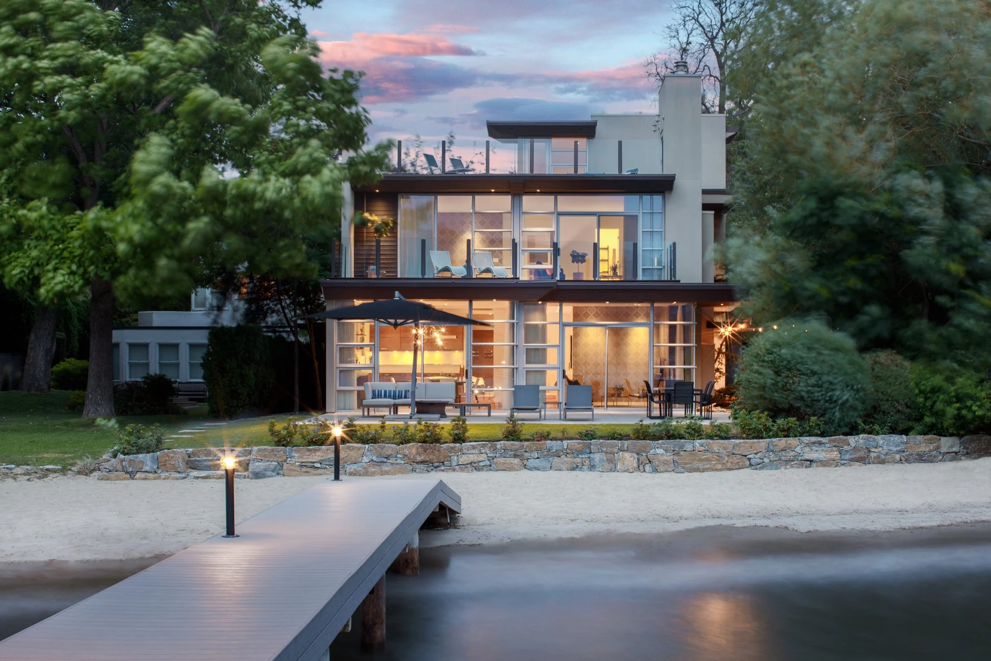 Lakefront house with large glass windows and outdoor patio, illuminated inside at dusk, surrounded by trees, with a wooden walkway leading to water with sandy shore.