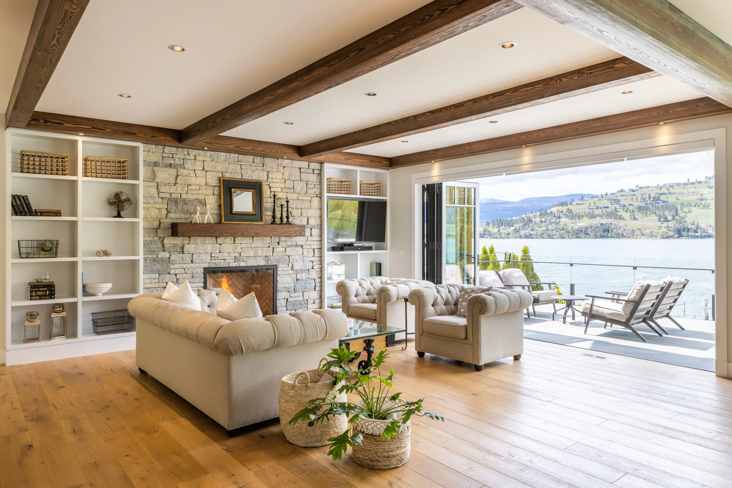 Waterfront home in Okanagan Landing, with a living room with ceiling beams, stone fireplace, white bookshelf, beige sofas, and large window overlooking a lake and hills.