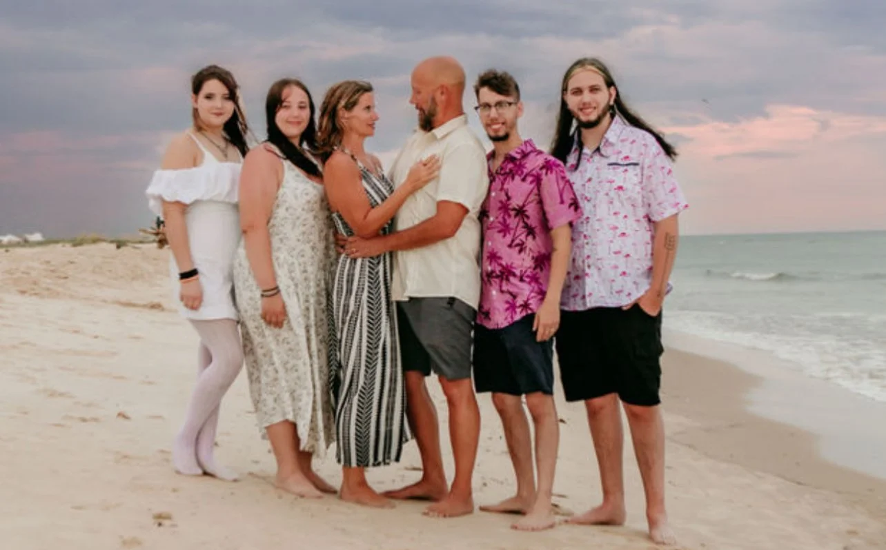 A group of six people standing on a sandy beach during sunset, facing each other and smiling, with the ocean and sky in the background.