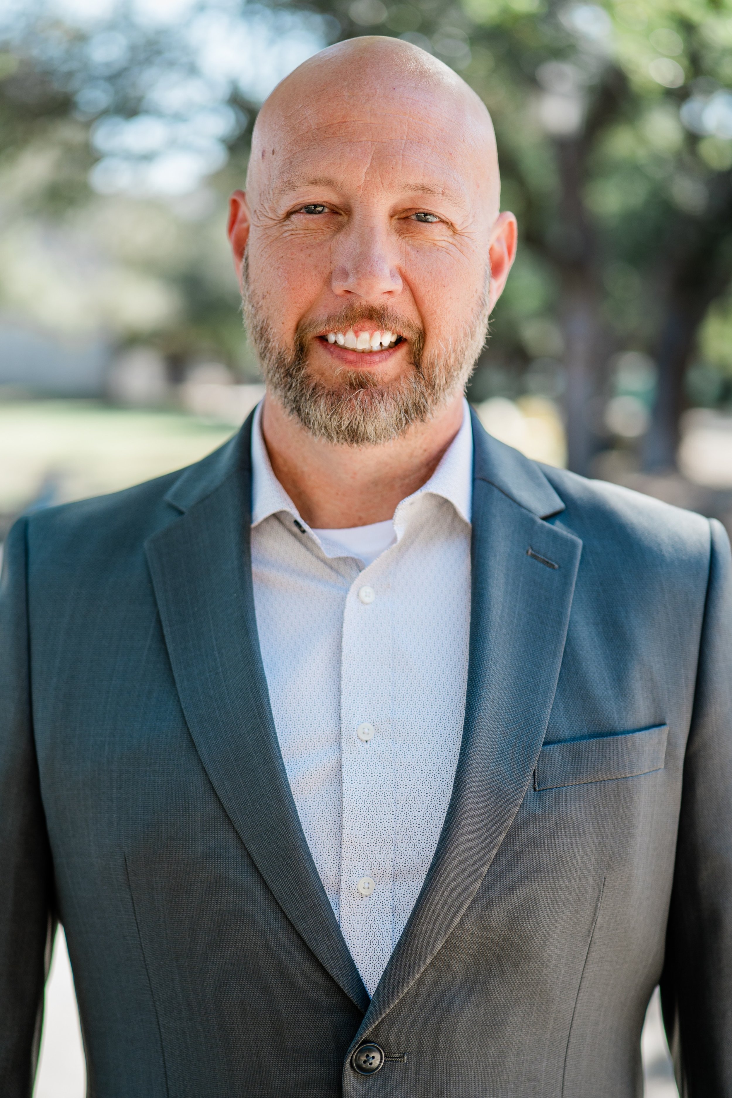 Close-up of a bald man with a beard, wearing a dark gray suit and white dress shirt, smiling, outdoors with greenery in the background.