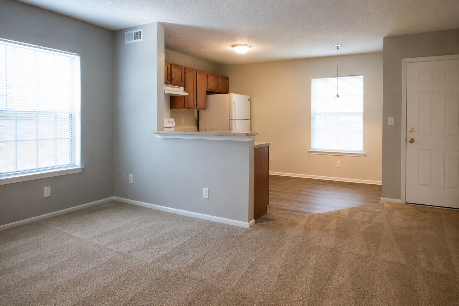 Empty living room with large windows, partially visible kitchen with wooden cabinets, refrigerator, and a small breakfast bar, and a front door in the background.
