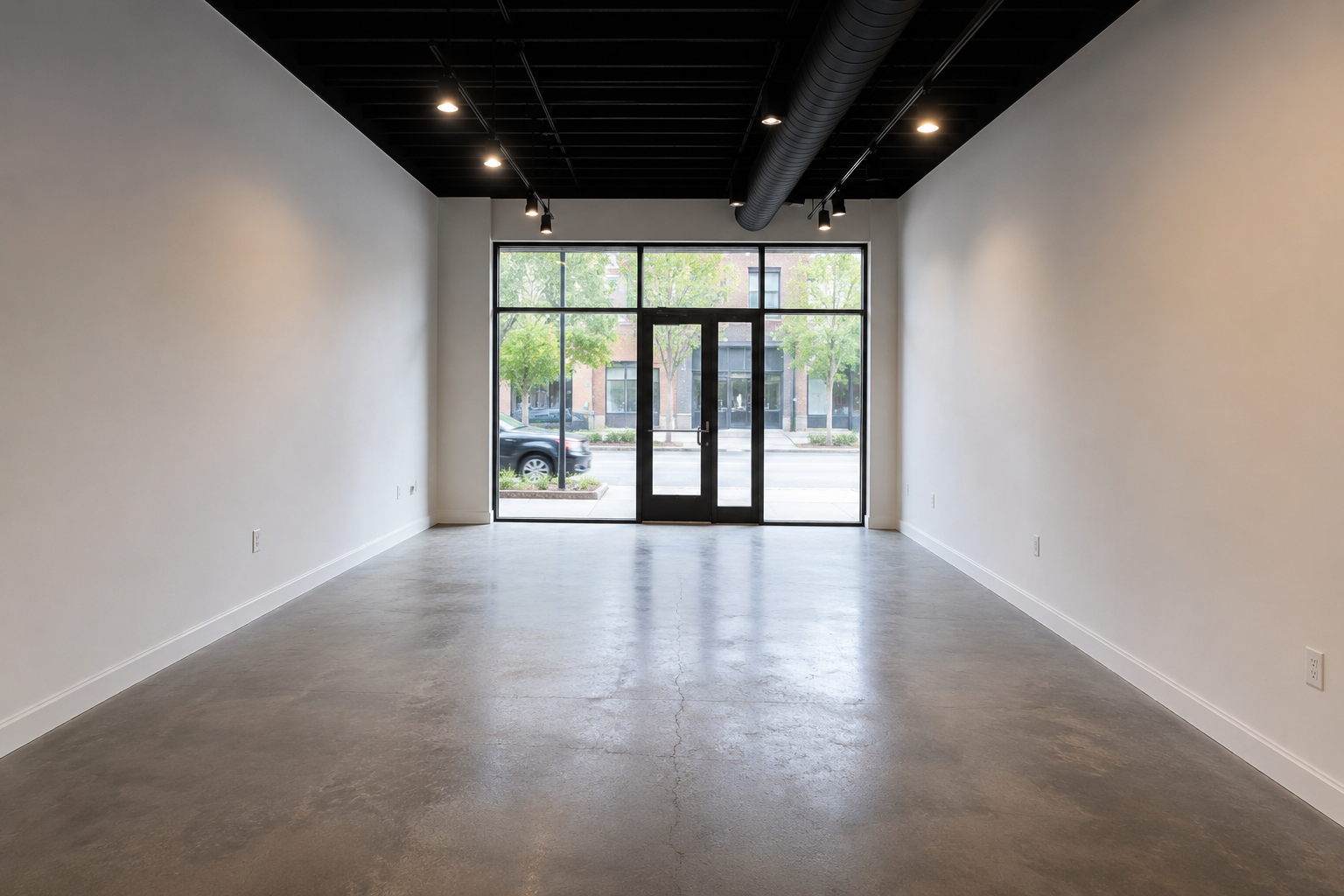 Empty commercial space with large front window and glass door, polished concrete floor, white walls, black ceiling with track lighting, and visible HVAC ductwork.