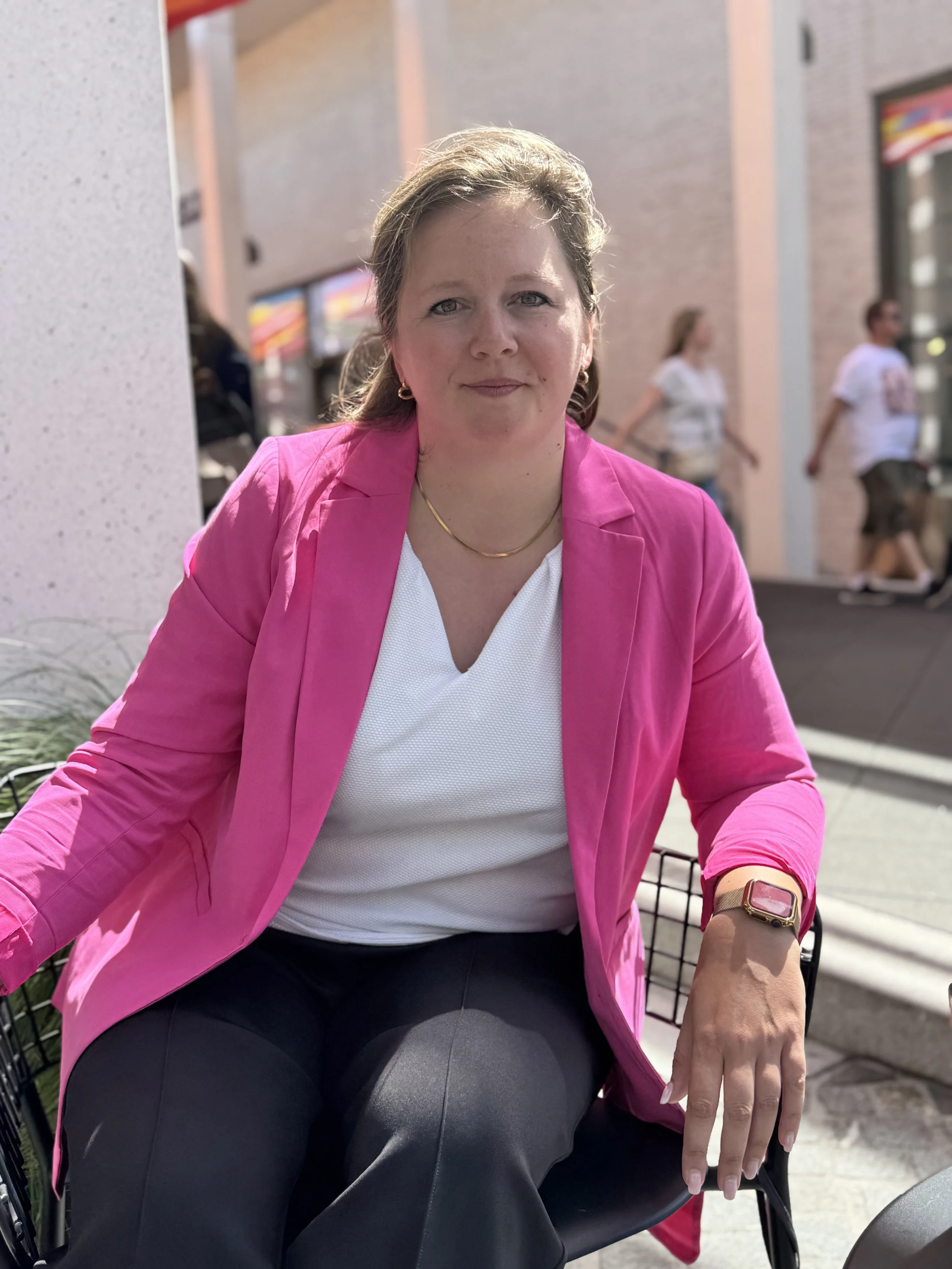 A woman in a pink blazer and white top sitting on a mobility scooter on a city sidewalk with people walking in the background.
