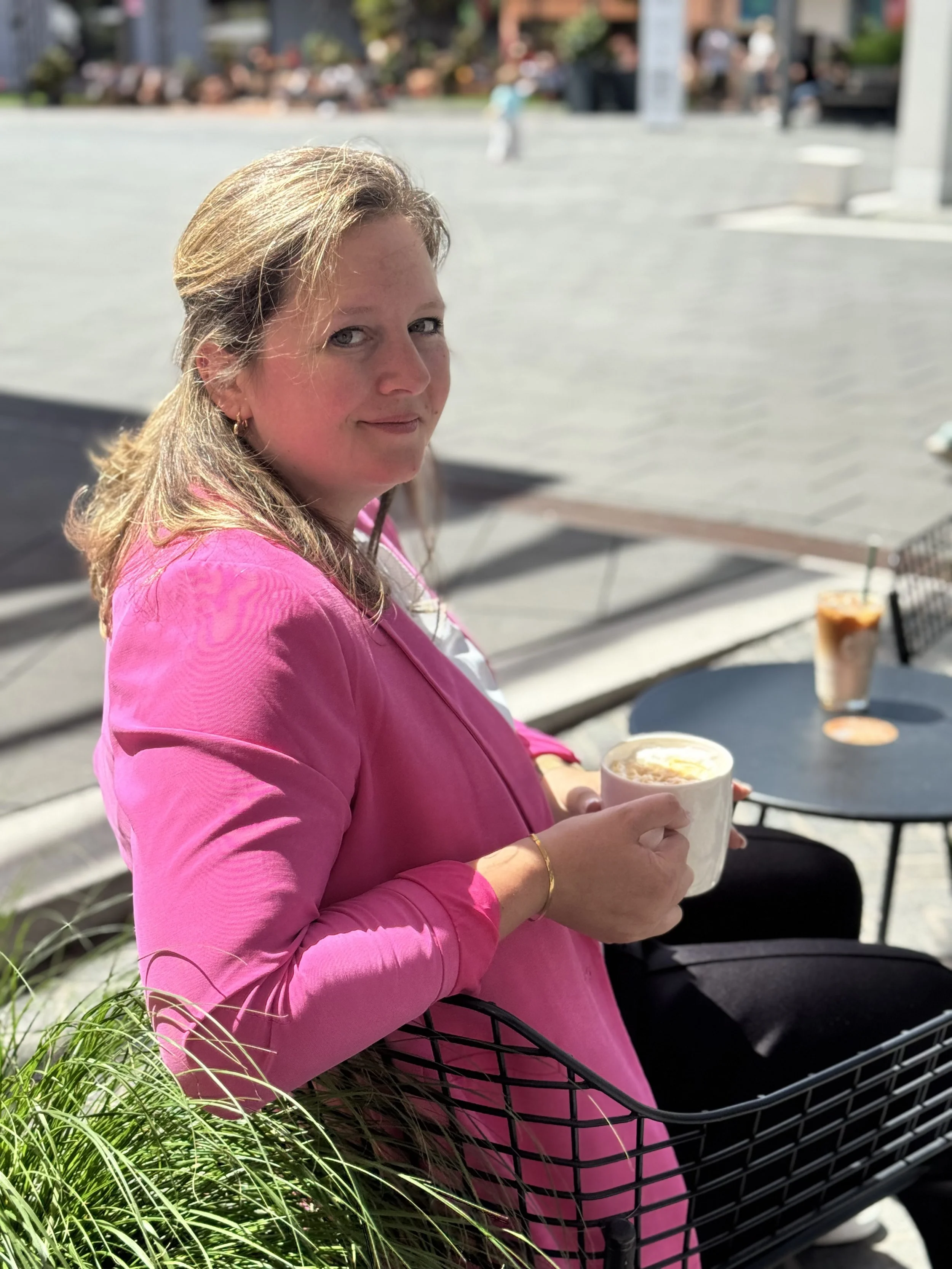 Woman with blonde hair wearing a pink jacket sitting outdoors in a shopping cart holding a drink, with blurred background of people and urban coaching environment.