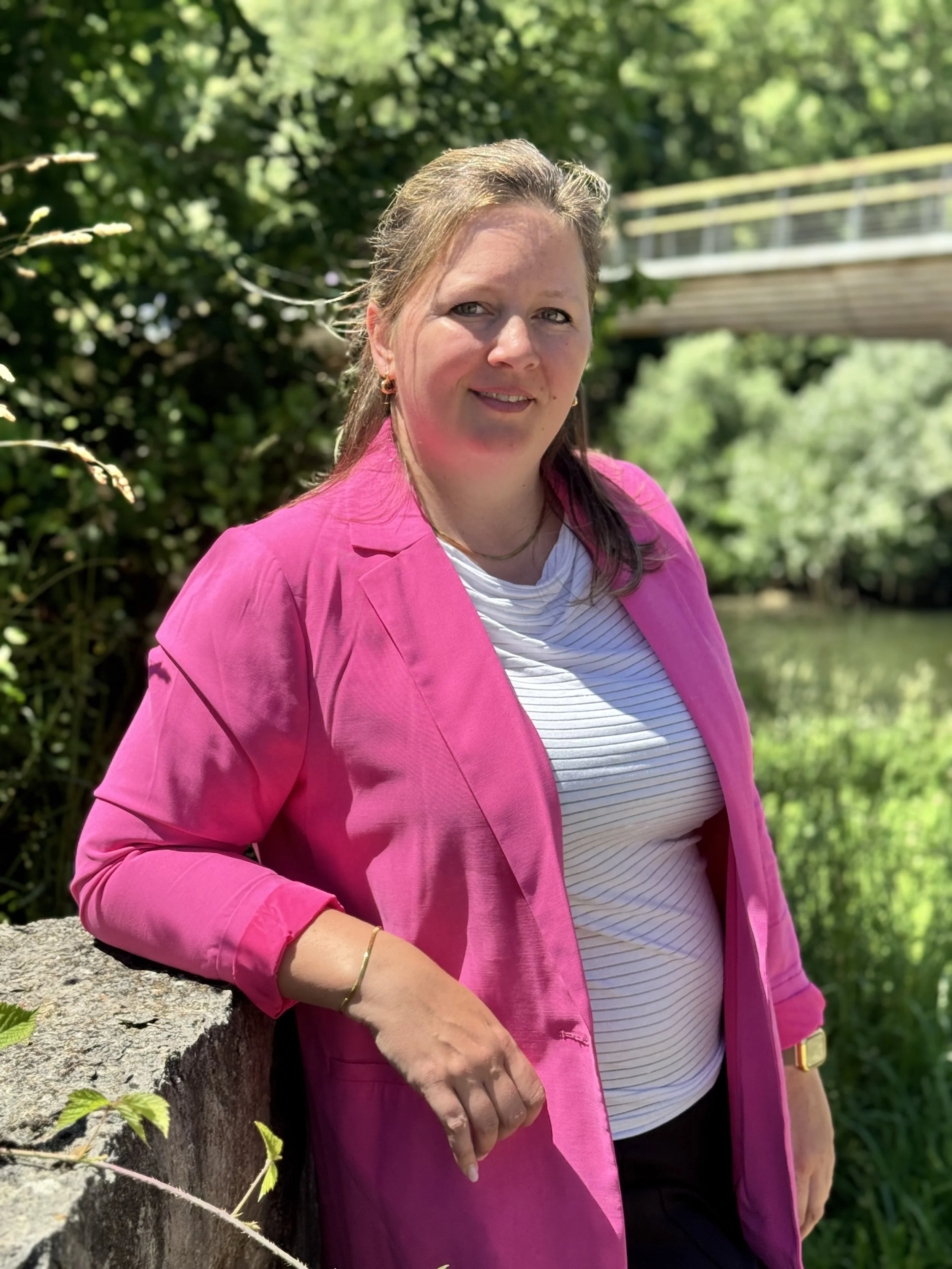 A woman in a pink blazer and white striped shirt leaning on a stone wall in an outdoor green park with a bridge in background.