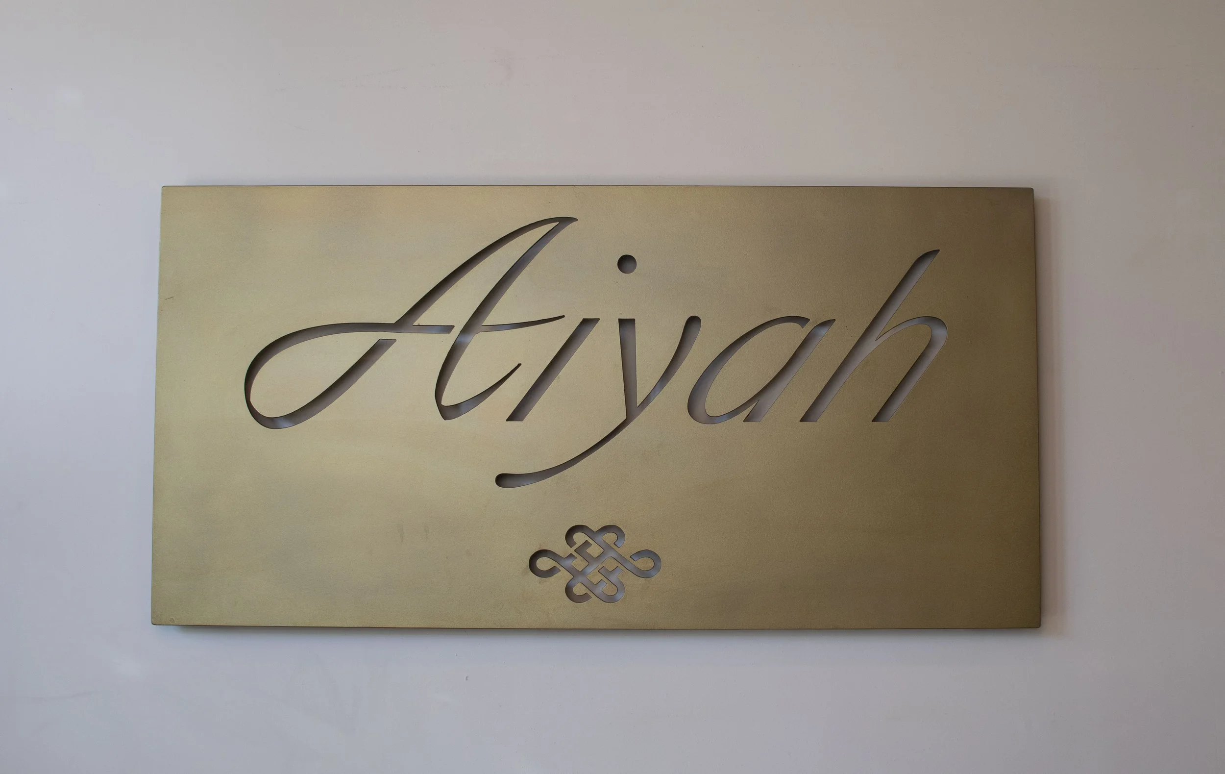 Gold-colored decorative wall plaque with the word 'faith' cut out in cursive script, featuring a decorative knot emblem below.